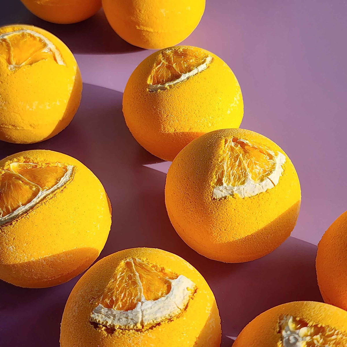 Bright yellow Energise aromatherapy bath bombs topped with dried orange slices arranged on a purple surface in natural light