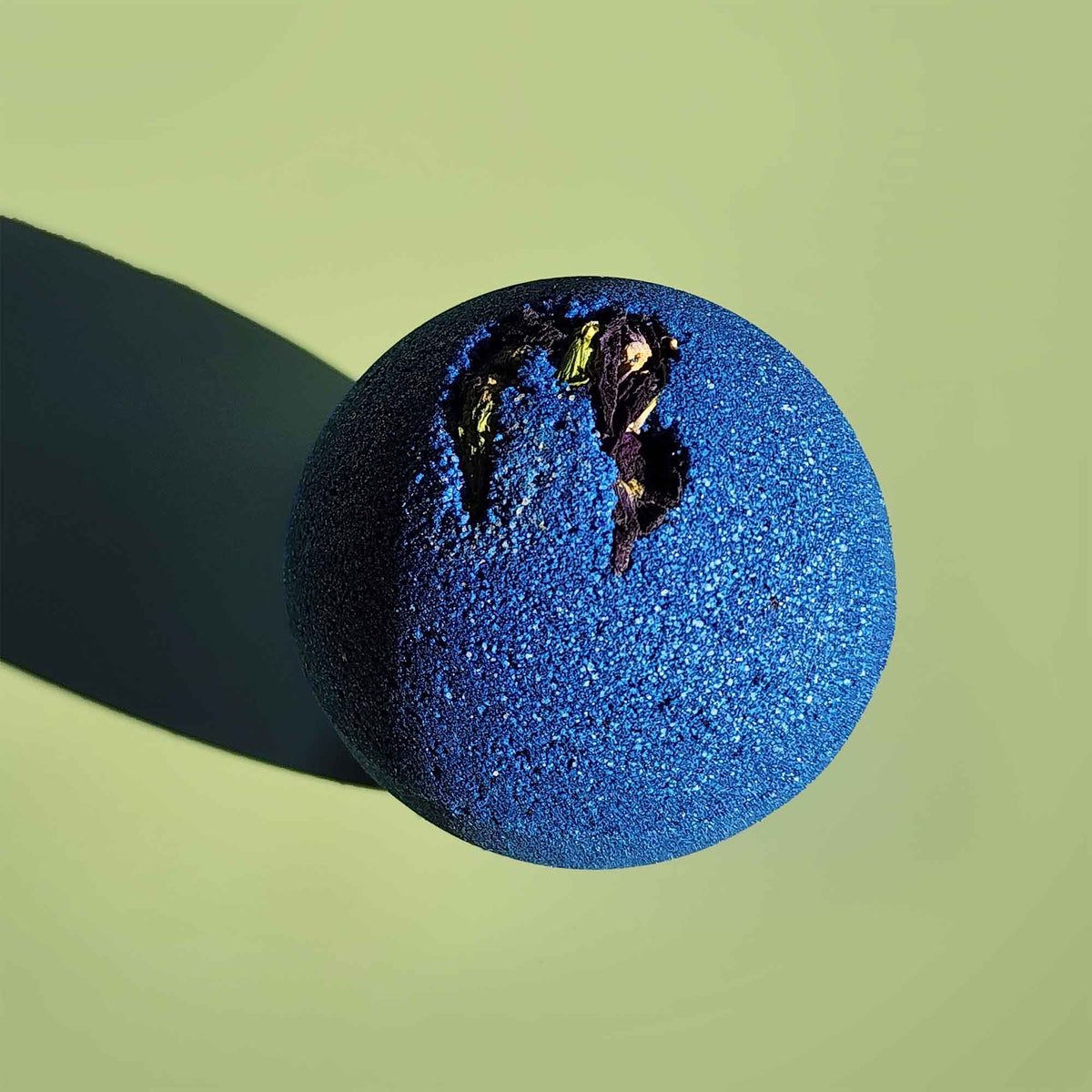 deep blue bath bomb with dried pea flowers on top on a plain green background