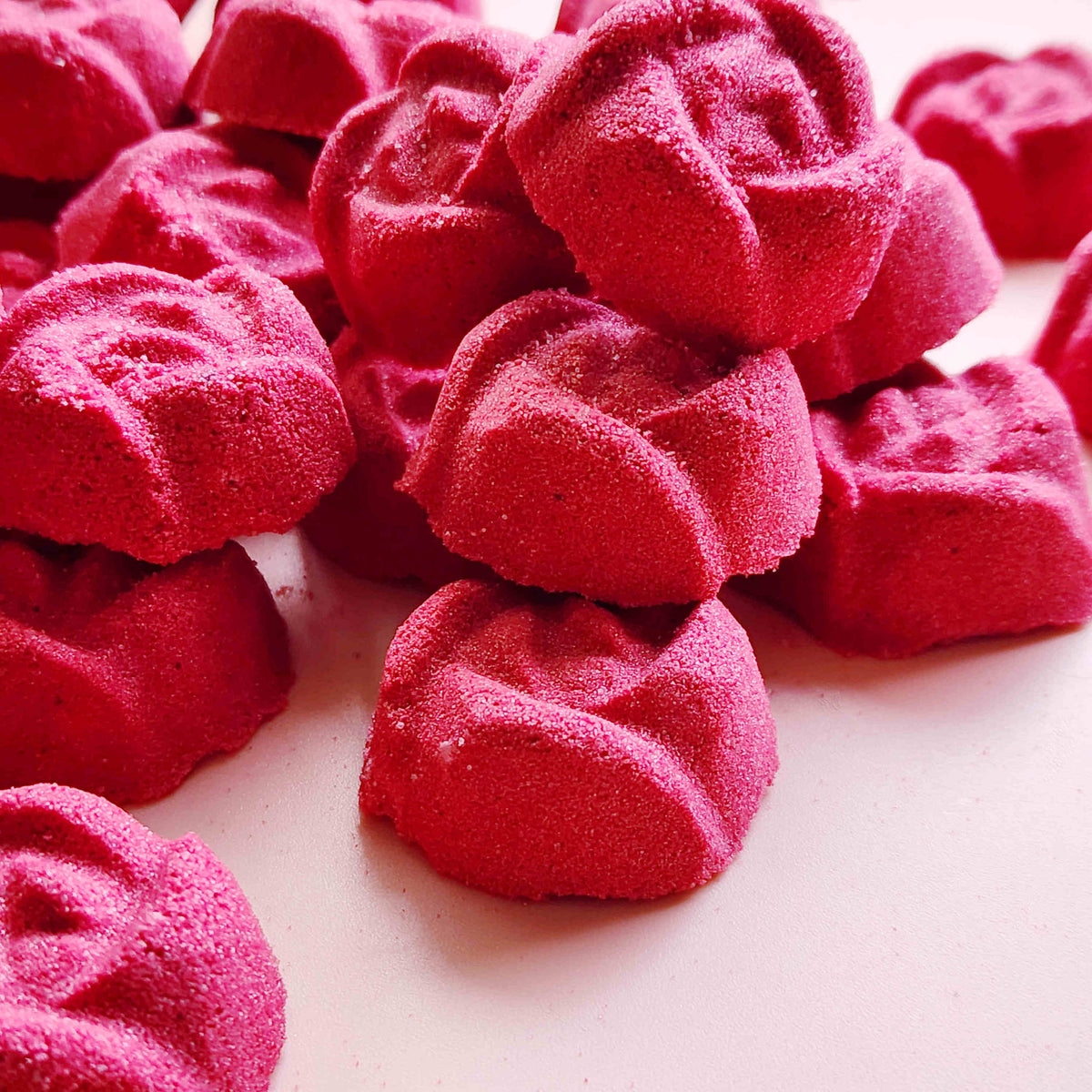 rose shaped mini bath bombs coloured a deep red laid out neatly on a plain pink background