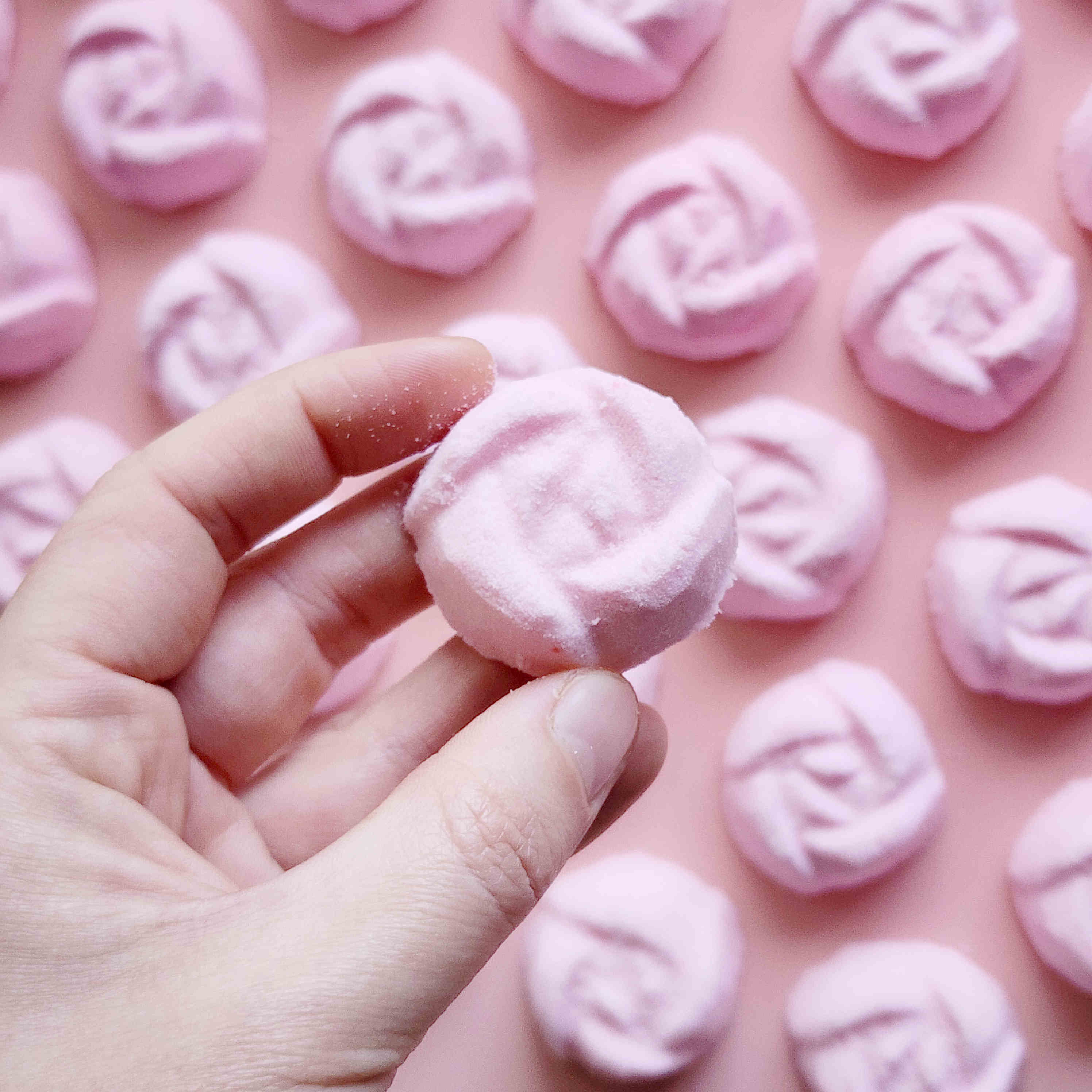 light dusty pink coloured mini bath bombs in the shape of roses on a pink background with one being held in a hand