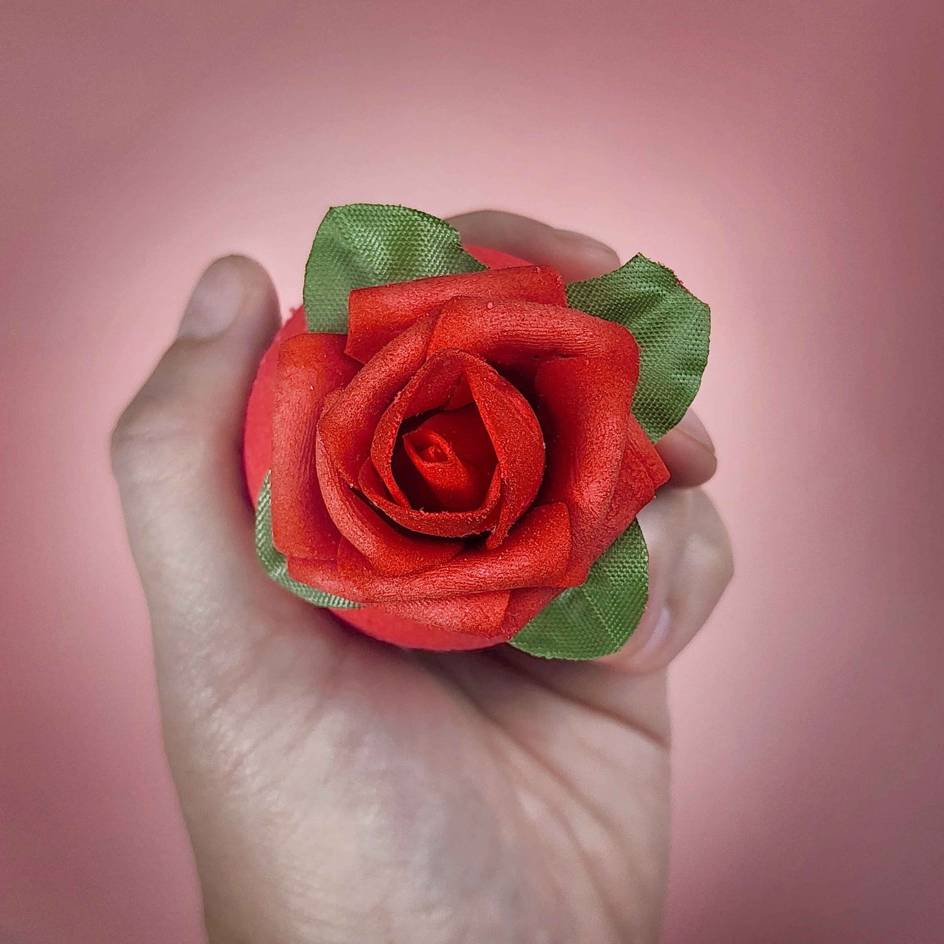 Red circular bath bomb with a rose on top held in a hand on a pink backdrop