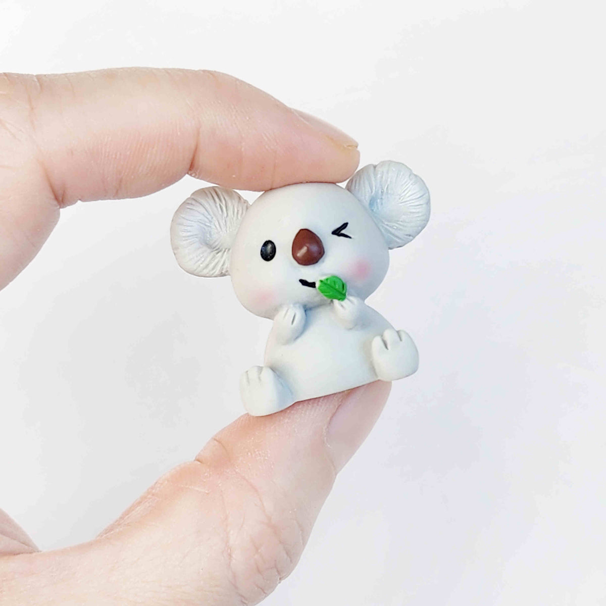 Cute grey koala resin figurine being held between two fingers on a plain white background