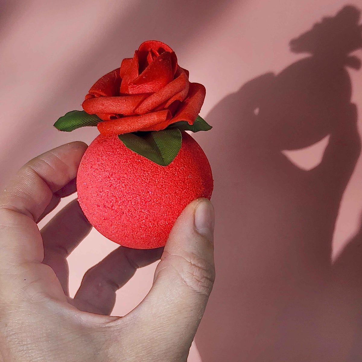 red round bath bomb with a foam rose on top being held in a hand on a pink background