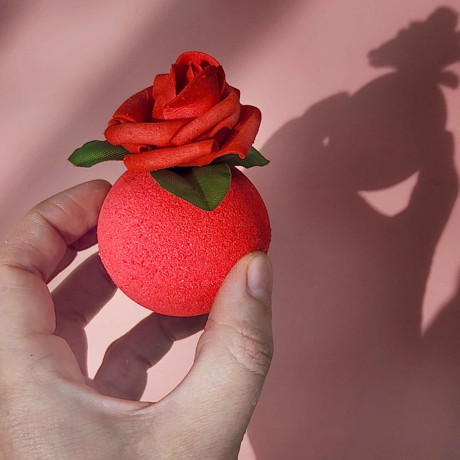 Red circular bath bomb with a rose on top held in a hand on a pink backdrop