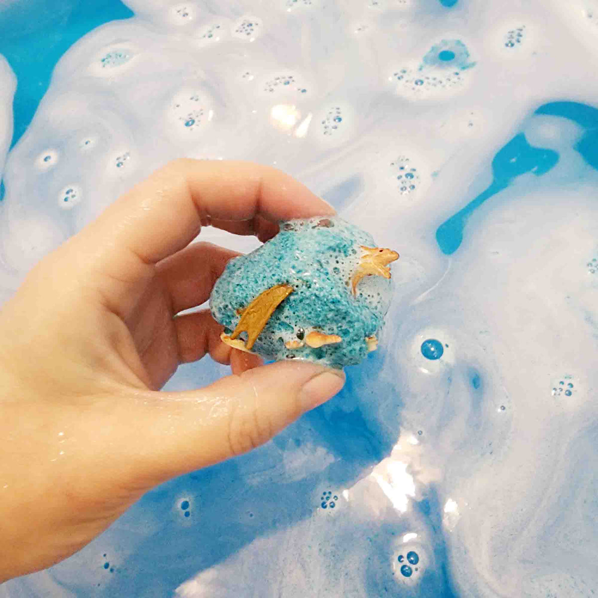 dragon toy partially revealed in a semi dissolved teal bath bomb in bright blue coloured water