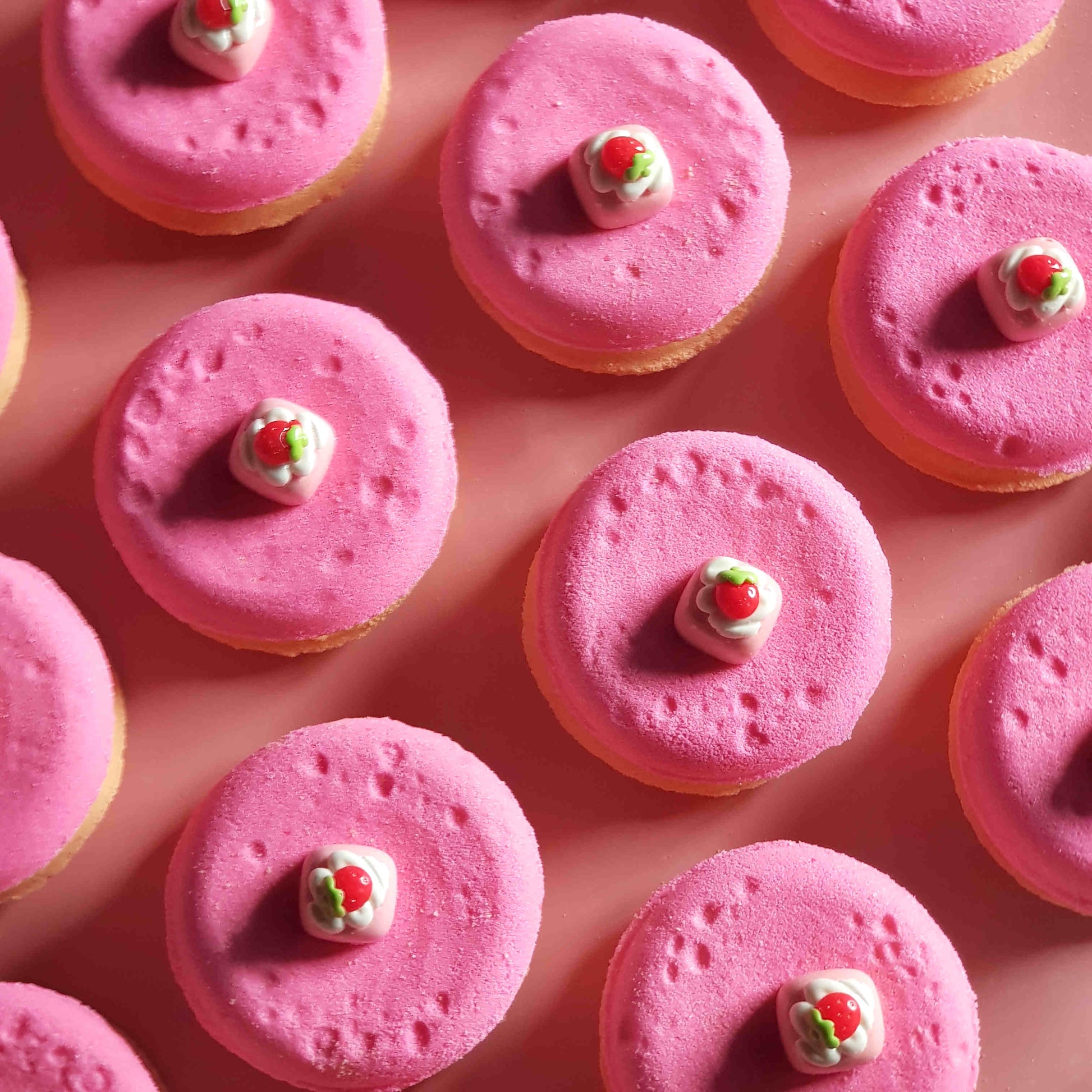 pink and beige disc shaped bath bombs with small strawberry dessert charms on top placed on a plain pink background