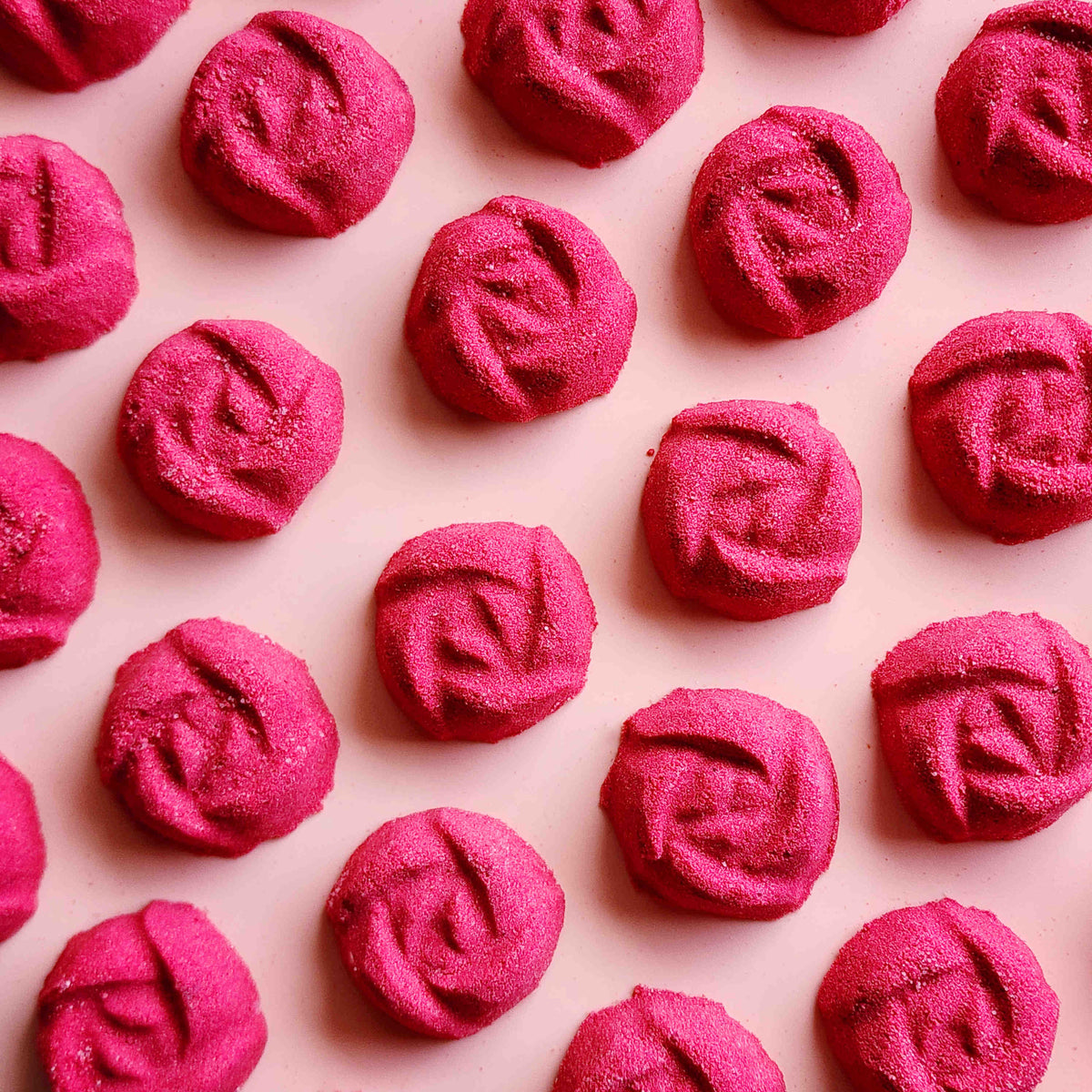 rose shaped mini bath bombs coloured a deep red laid out neatly on a plain pink background