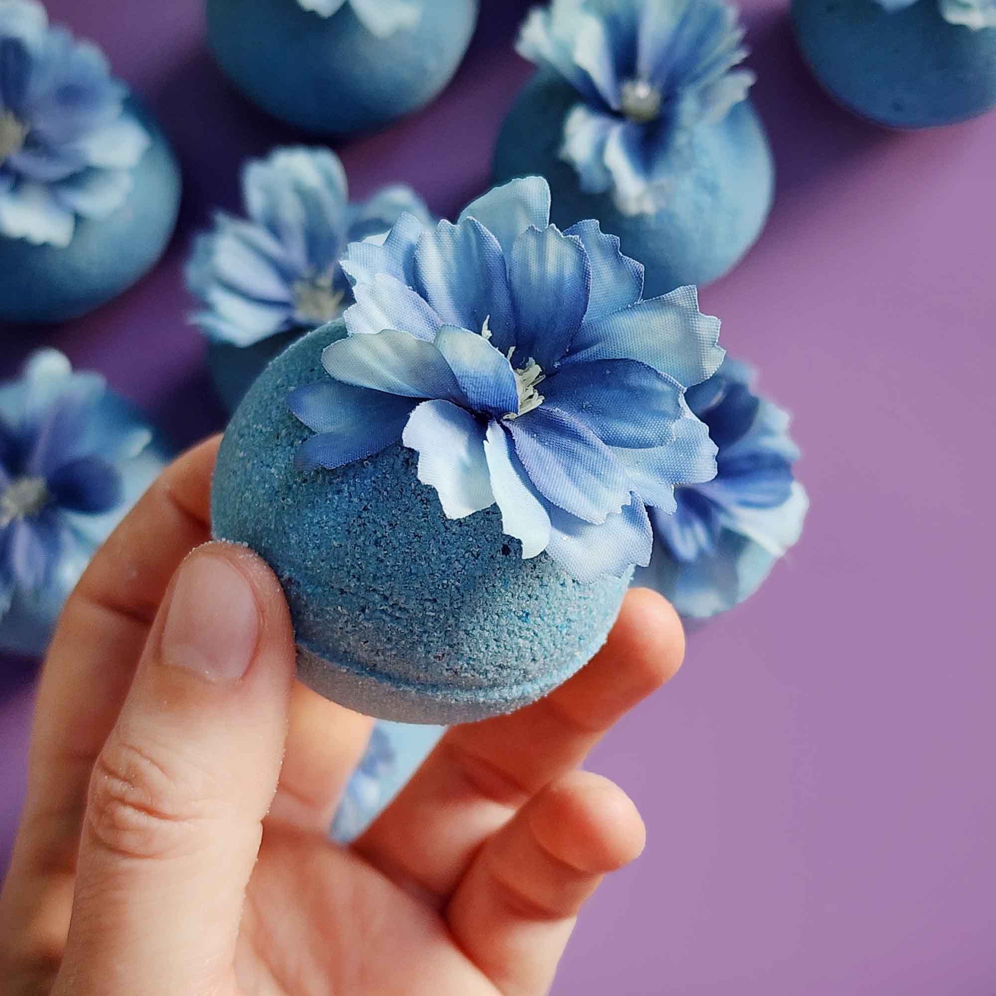 Hand holding a single blue wildflower bath bomb topped with a delicate faux bloom, with more bath bombs blurred in the back
