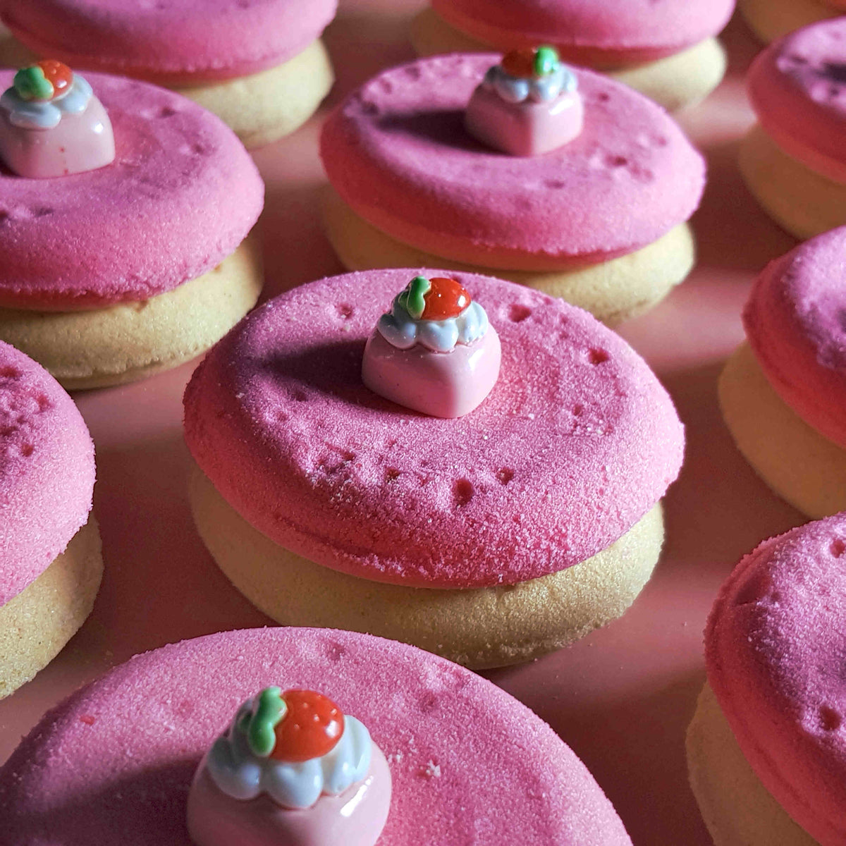 pink and beige disc shaped bath bombs with small strawberry dessert charms on top placed on a plain pink background