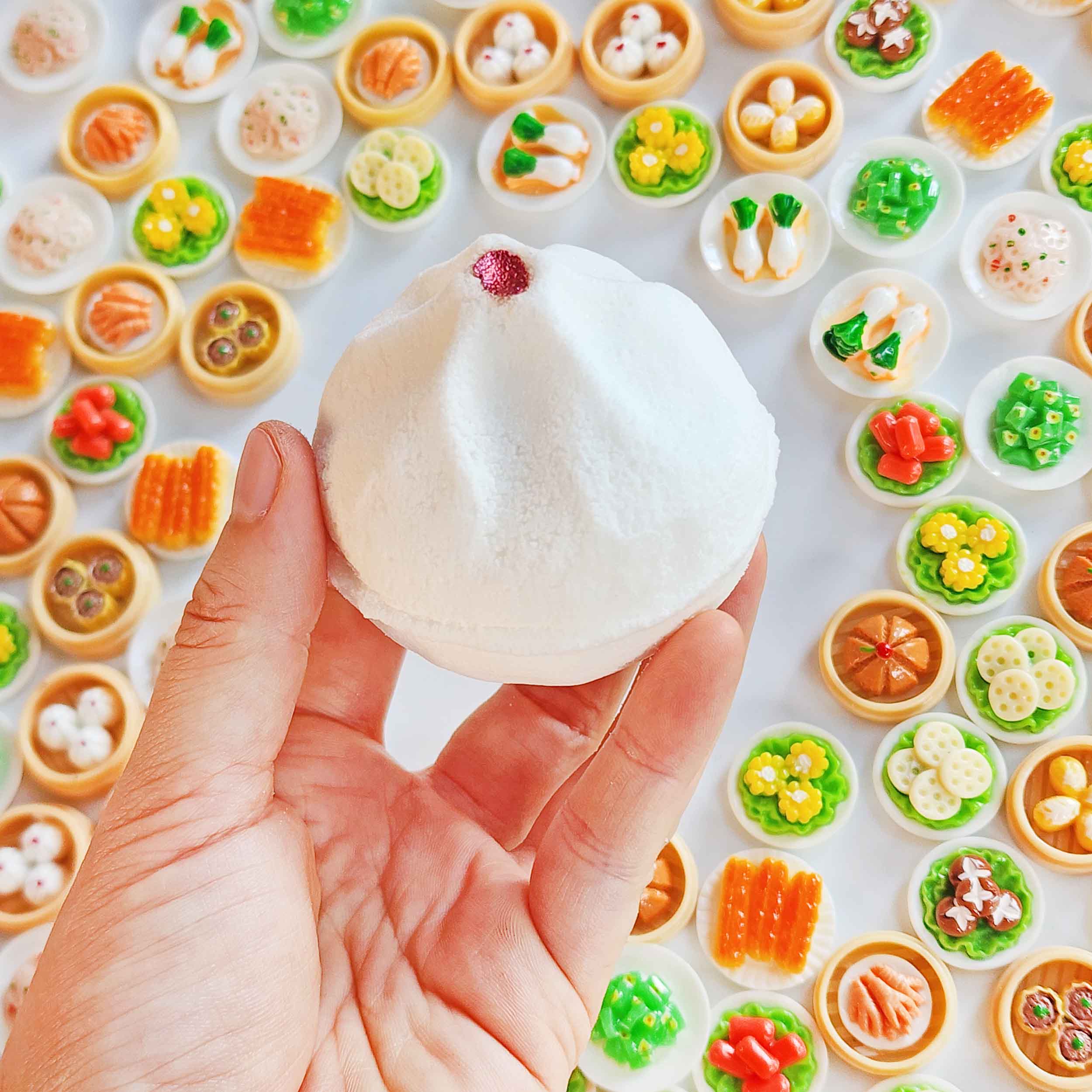 Hand holding a white Yum Cha bao bath bomb over miniature dim sum toys
