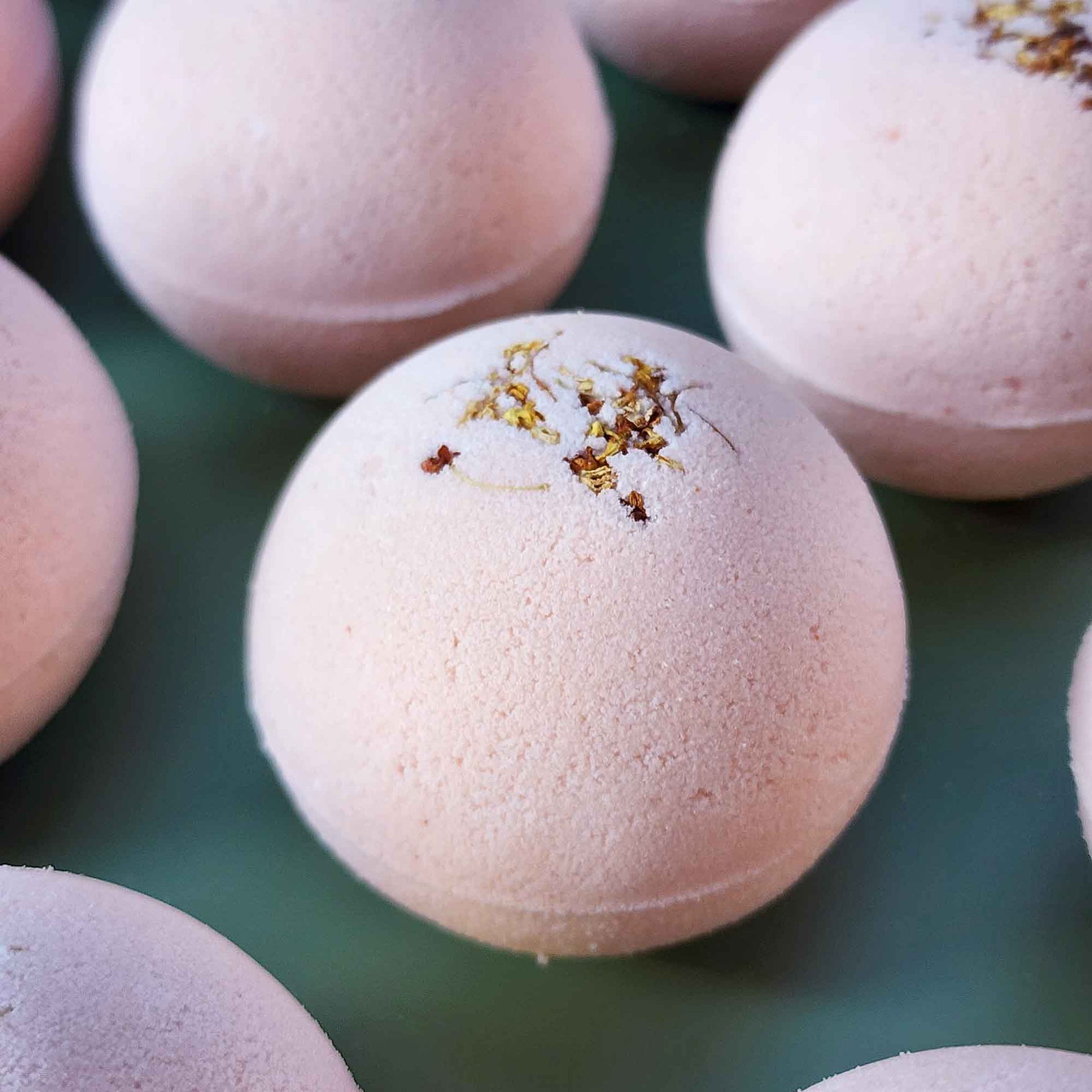 Multiple pale pink Grounding aromatherapy bath bombs topped with small dried osmanthus flowers, arranged on a soft green back