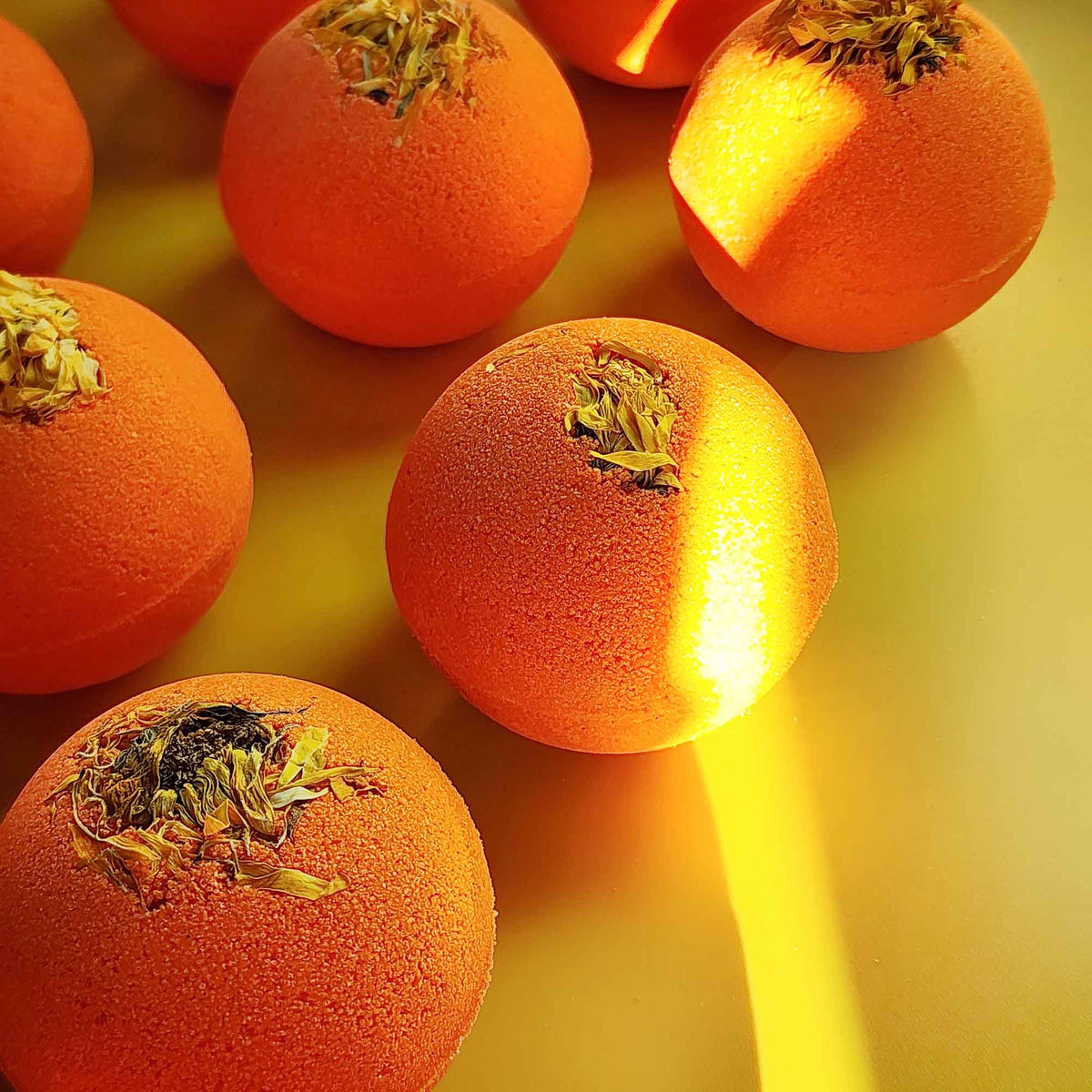 Bright orange Restore aromatherapy bath bombs topped with whole dried chrysanthemum flowers on a yellow surface with sunlight