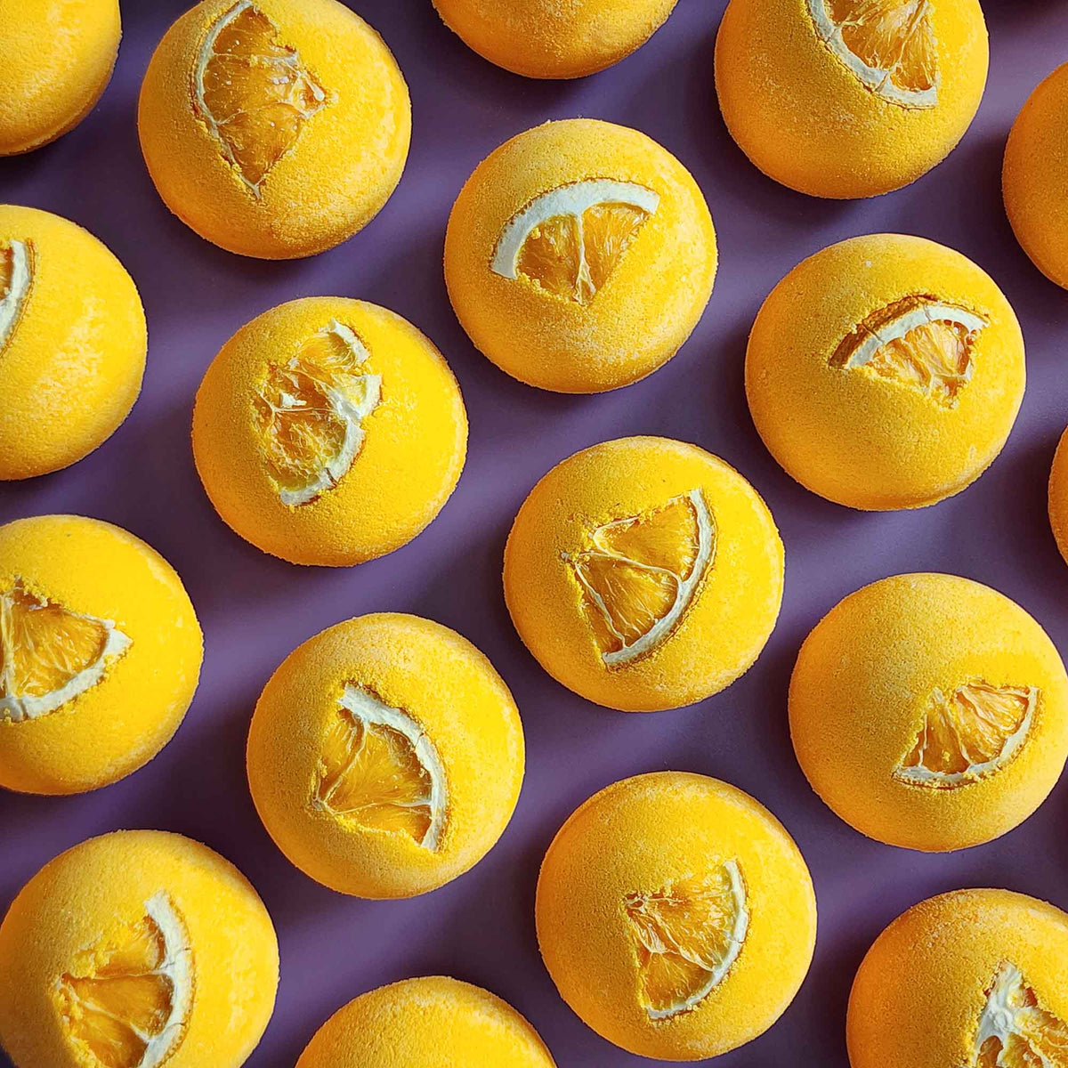Top-down view of multiple yellow Energise aromatherapy bath bombs topped with dried orange slices on a purple background