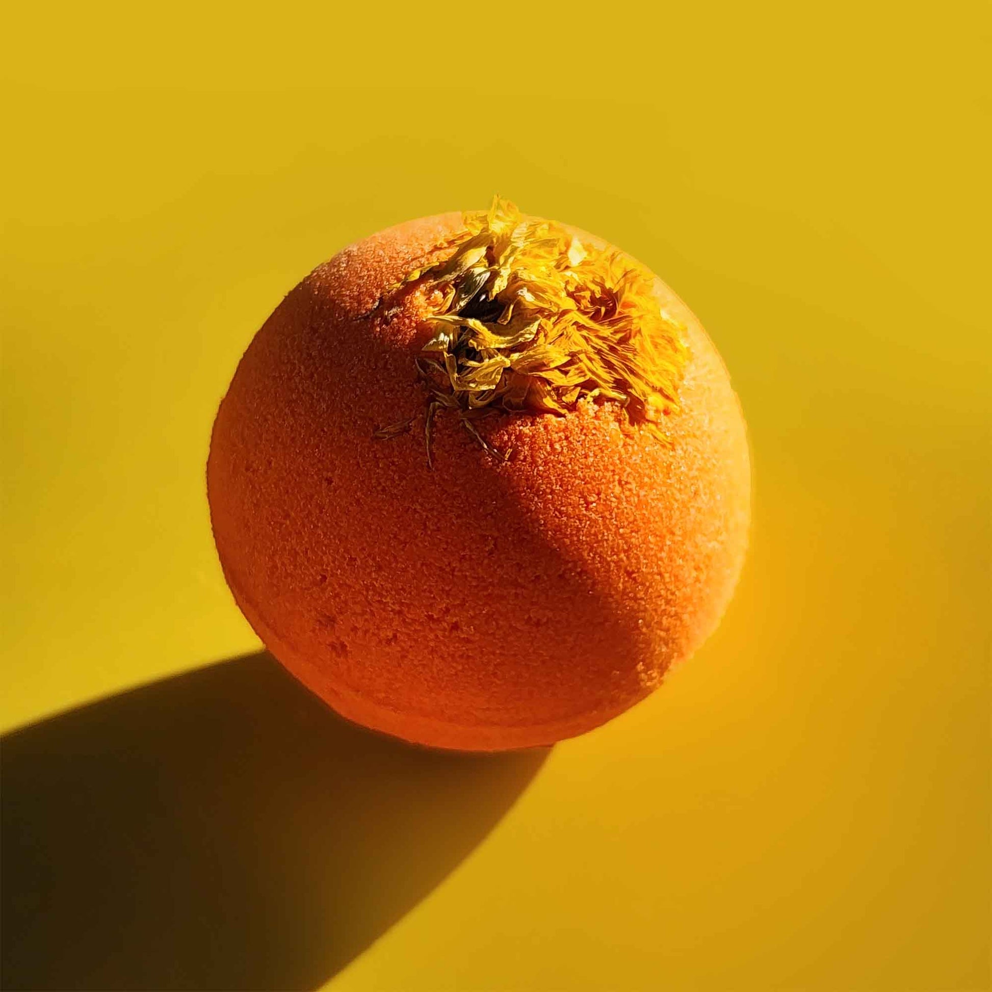 Close-up of a bright orange Restore aromatherapy bath bomb with a whole chrysanthemum flower on a yellow surface.