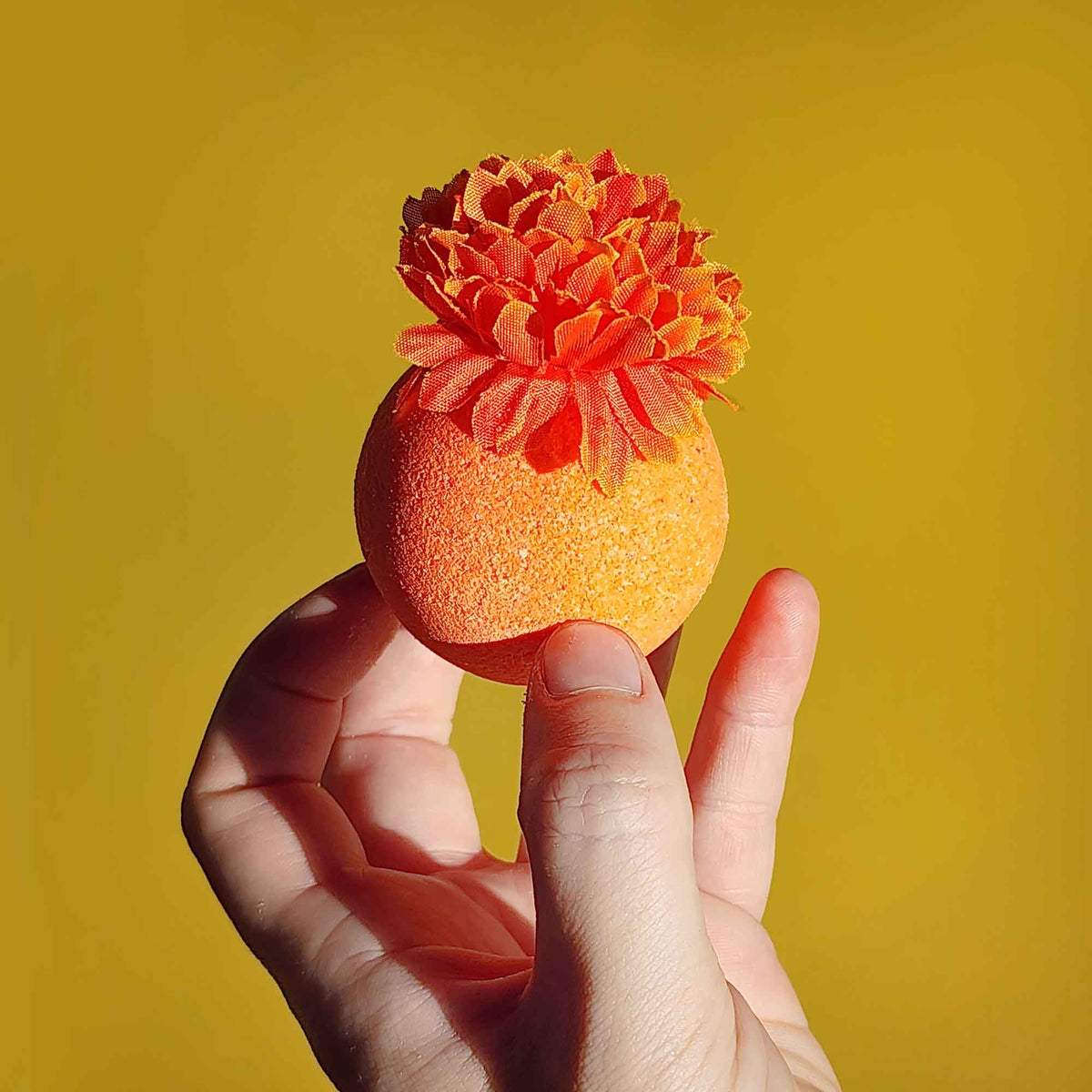 Hand holding a single orange bath bomb topped with a faux calendula flower against a yellow backdrop