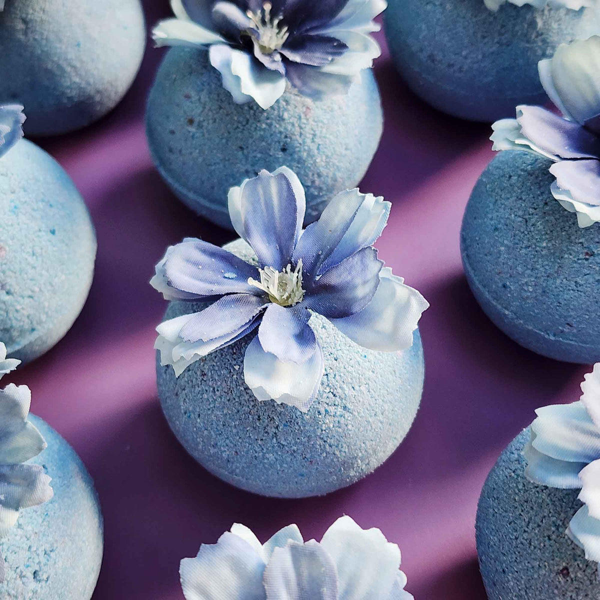 Close-up of handmade wildflower-scented bath bombs in dusty blue tones, each topped with blue faux petals on mauve background