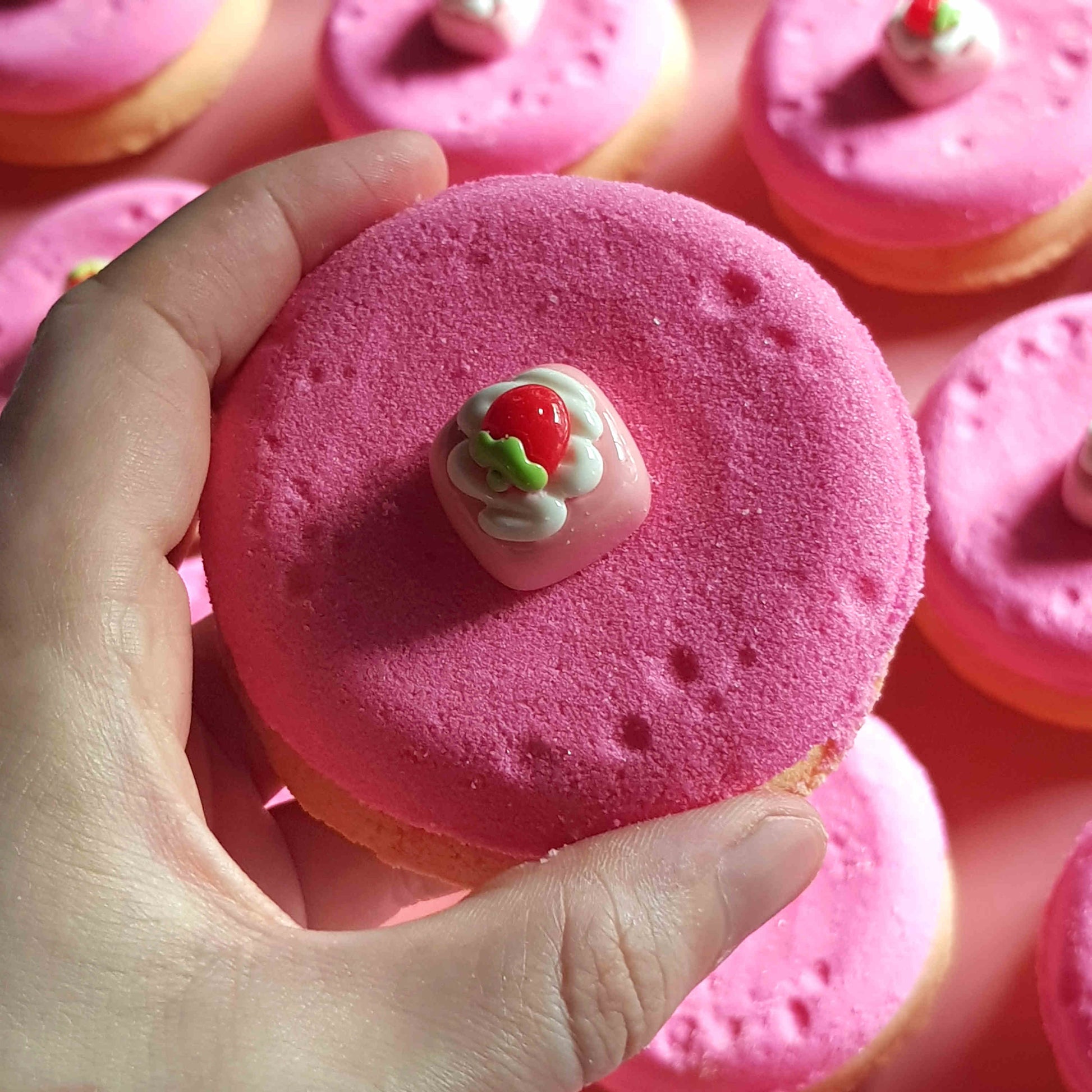 pink and beige disc shaped bath bombs with small strawberry dessert charms on top being held in a hand