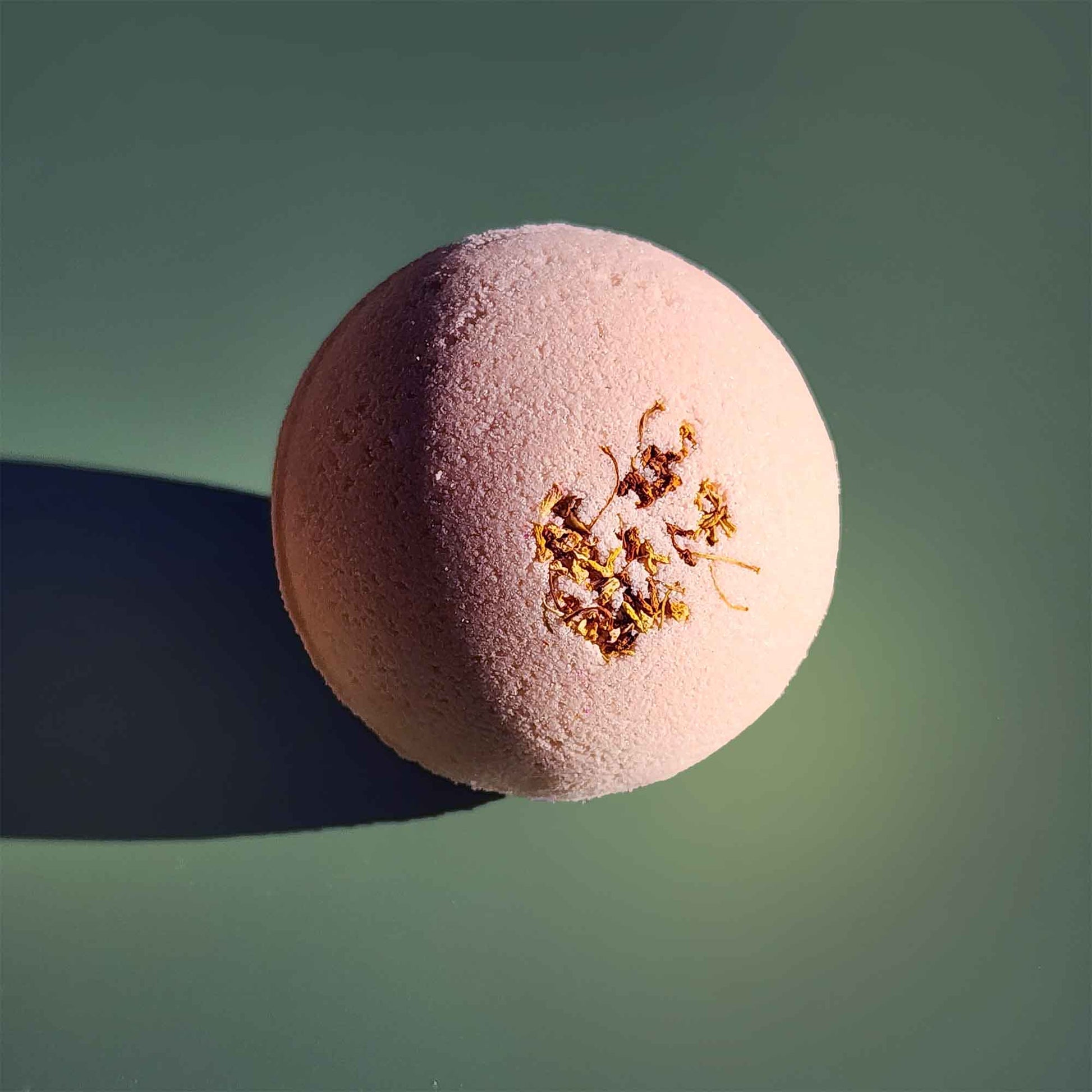 Close-up of a pale pink Grounding aromatherapy bath bomb topped with small dried osmanthus flowers on a soft green background