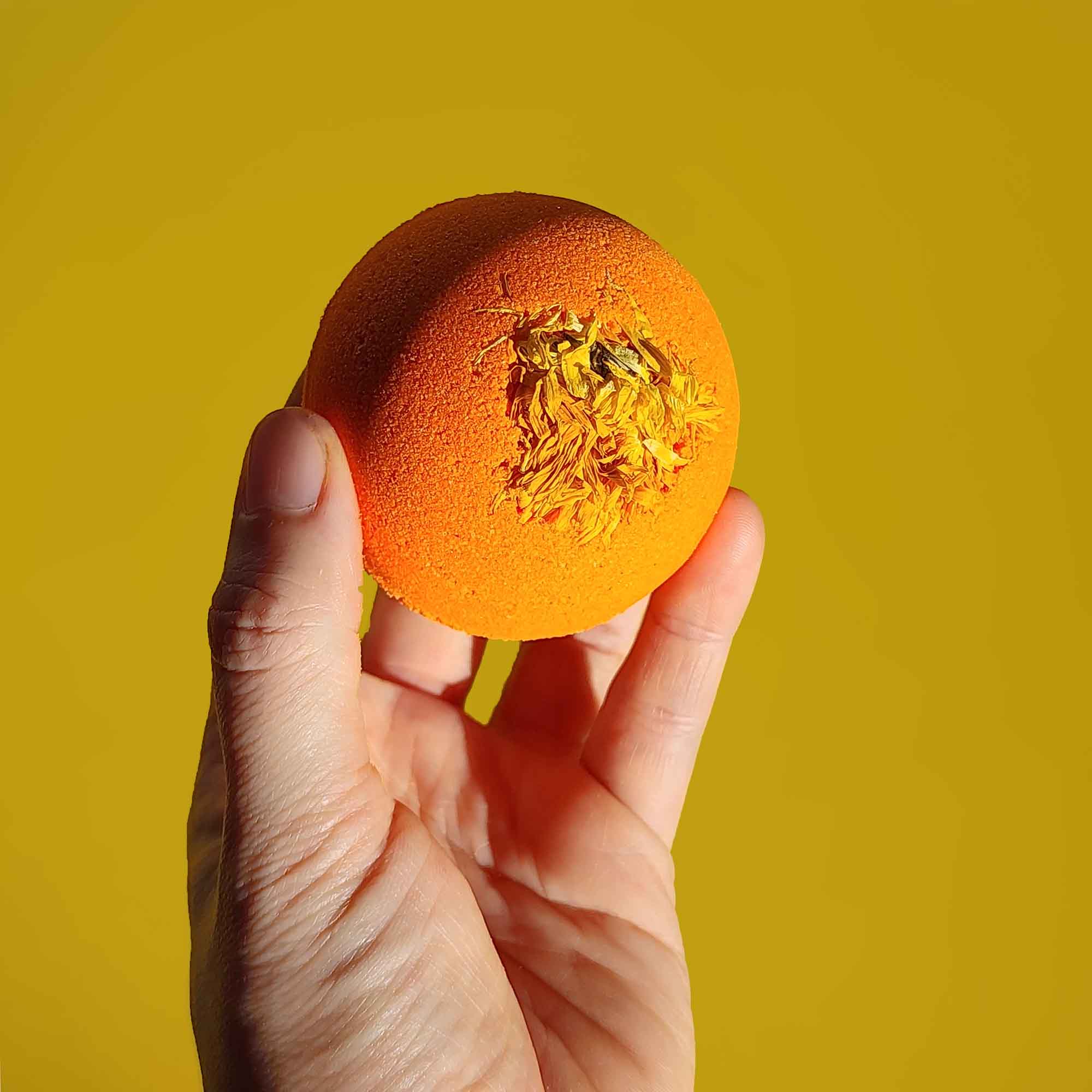 A hand holding a bright orange Restore aromatherapy bath bomb topped with a chrysanthemum flower against a yellow background