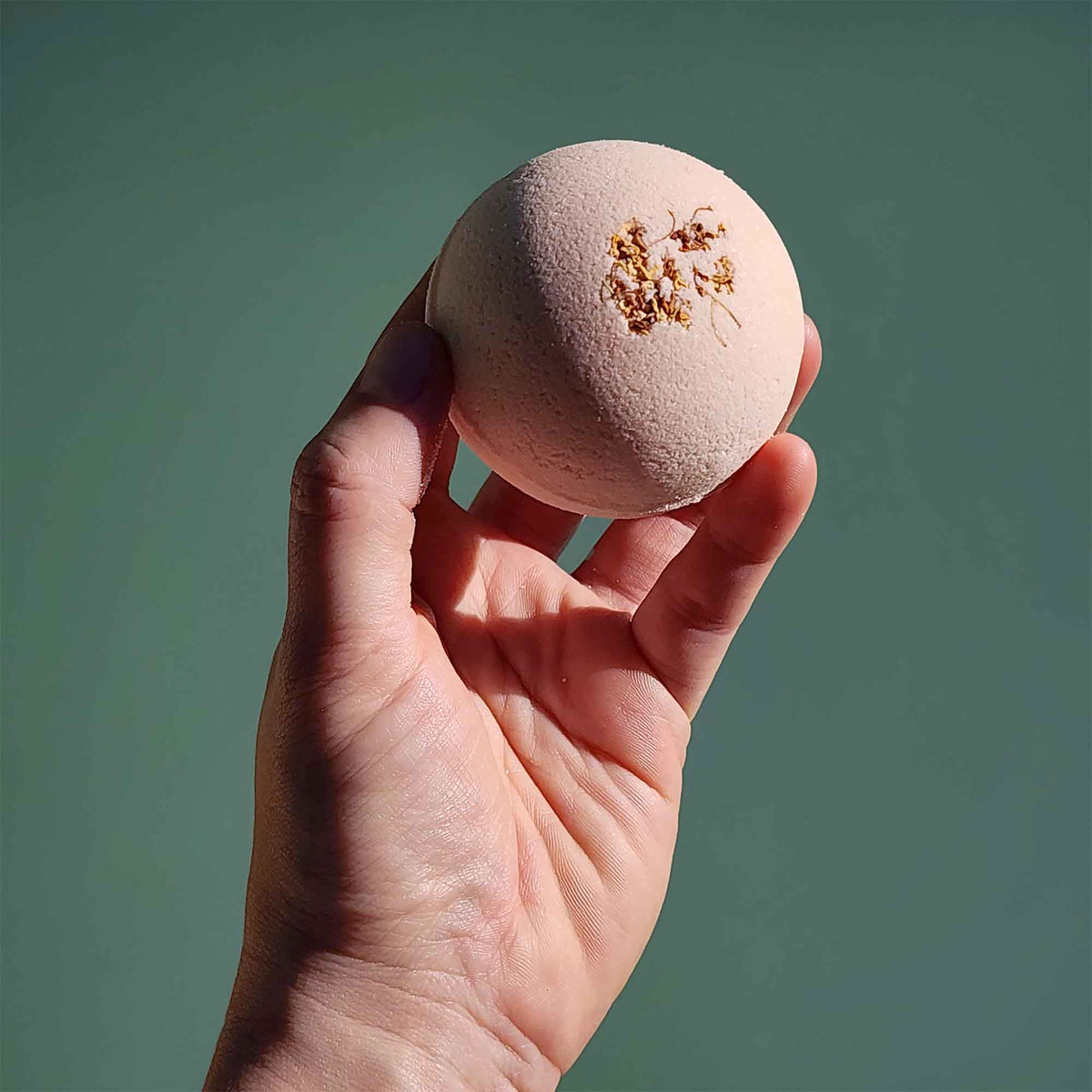 A hand holding a pale pink Grounding aromatherapy bath bomb topped with dried osmanthus flowers against a soft green backgrou