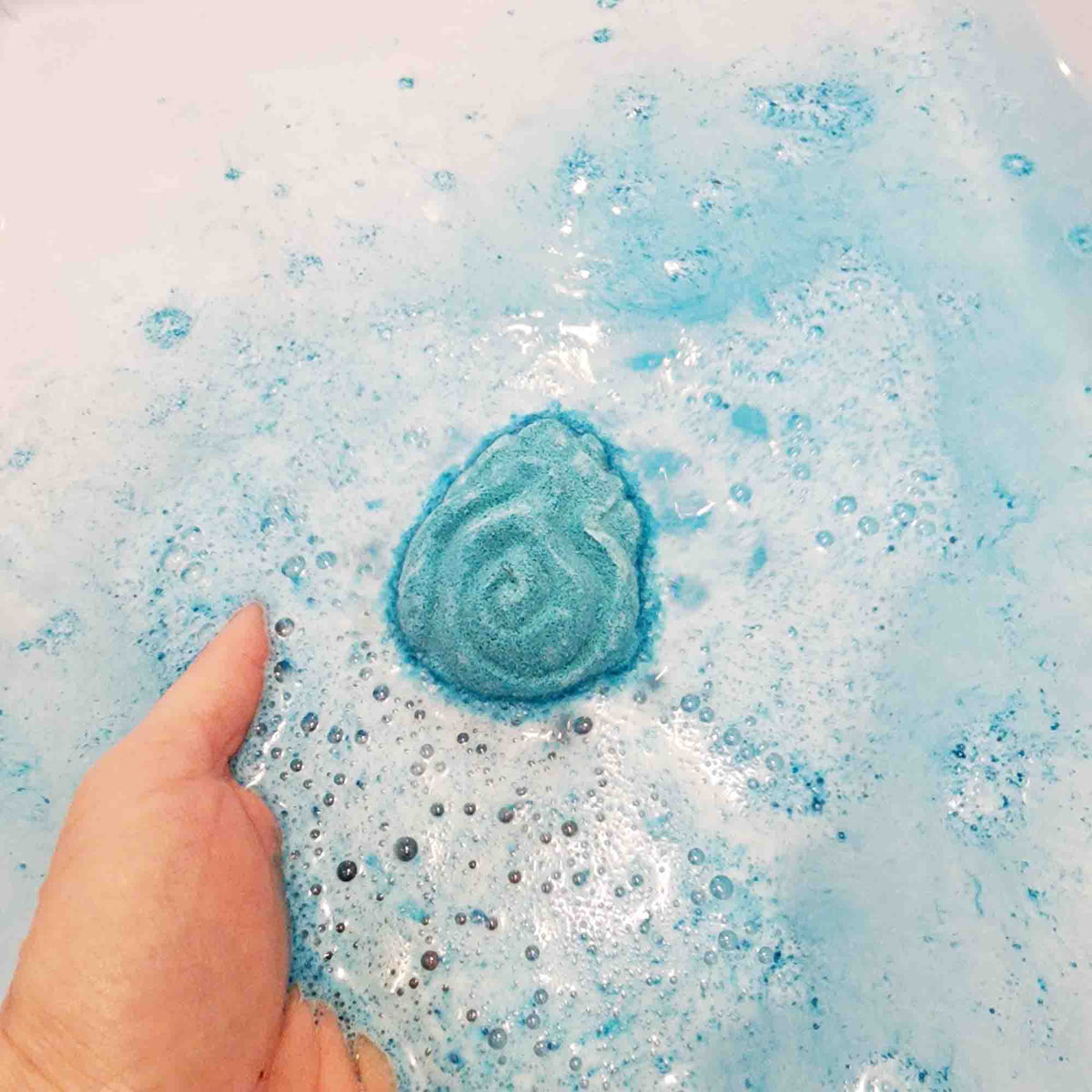 bright teal egg shaped bath bomb with intricate carvings in the shape of flames being held in the water surrounded by bubbles