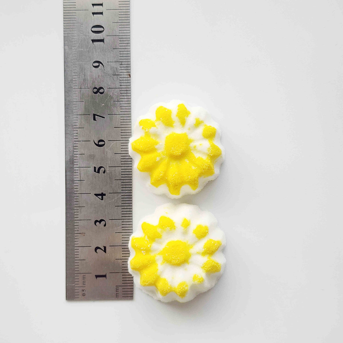 chamomile shaped mini bath bombs with a white base and yellow detail on a neutral plain background with a ruler