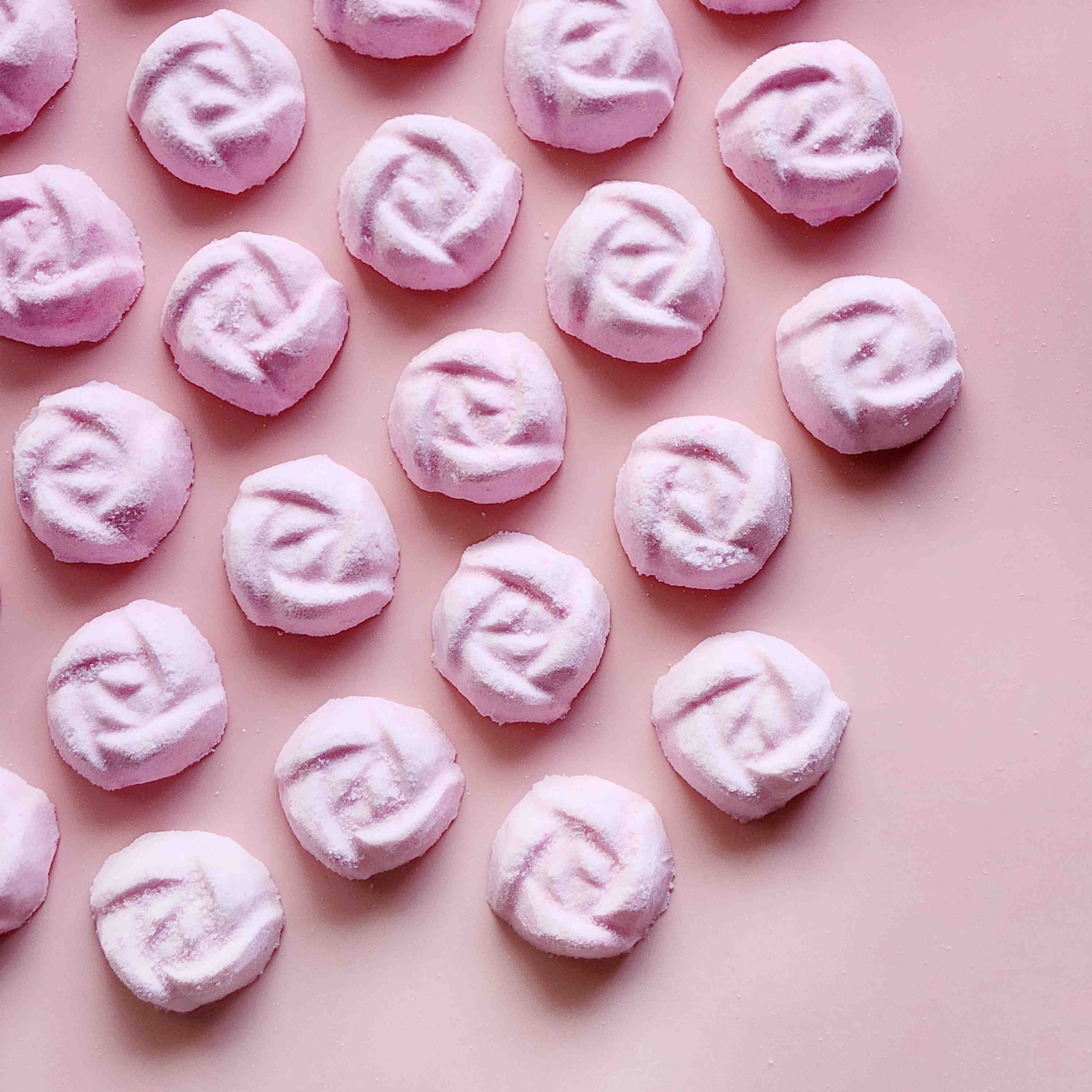 light dusty pink coloured mini bath bombs in the shape of roses laid out neatly on a plain pink background