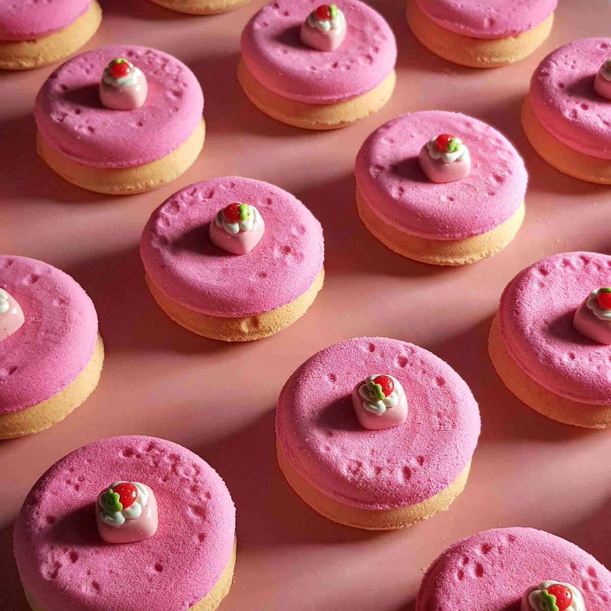 pink and beige disc shaped bath bombs with small strawberry dessert charms on top placed on a plain pink background