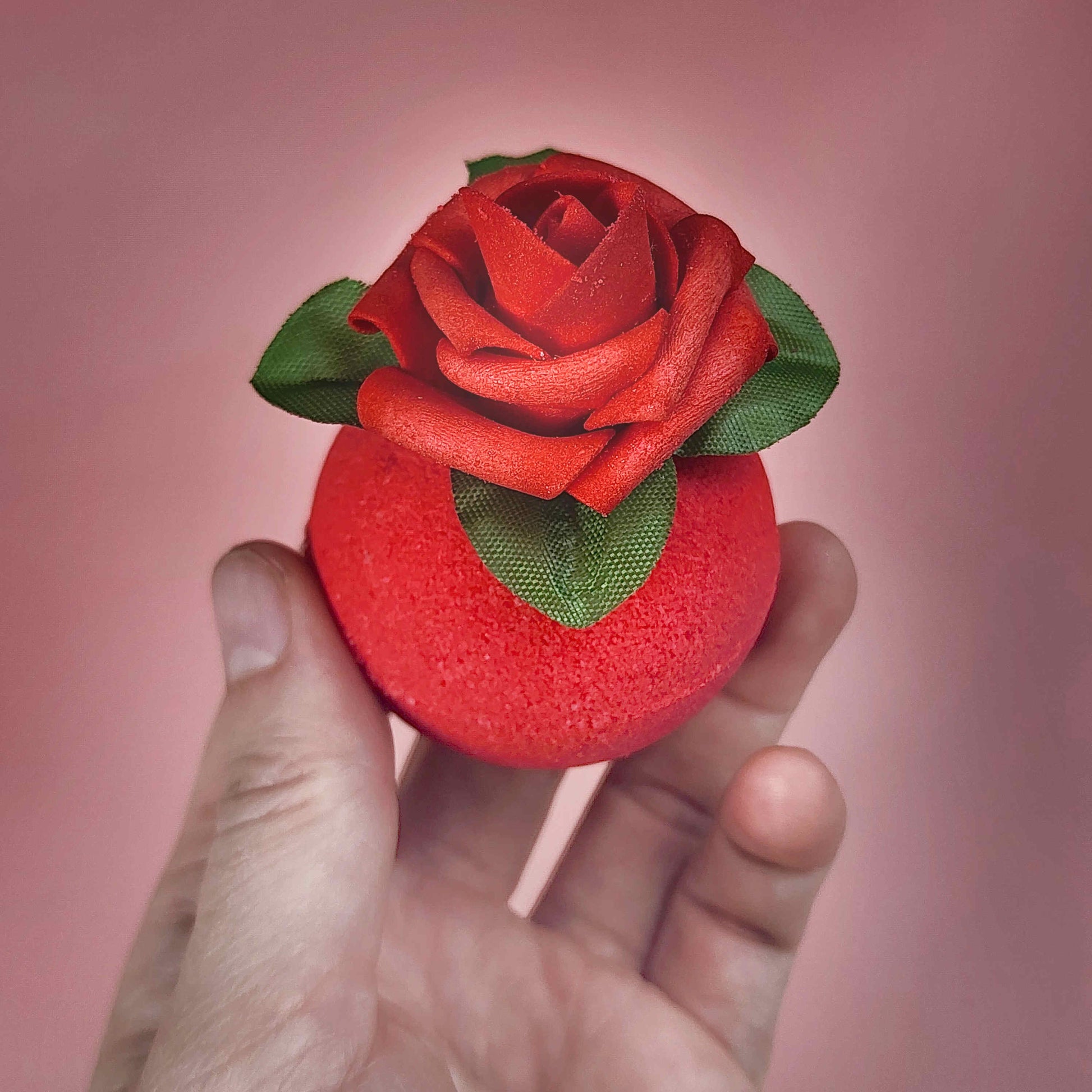Red circular bath bomb with a rose on top held in a hand on a pink backdrop