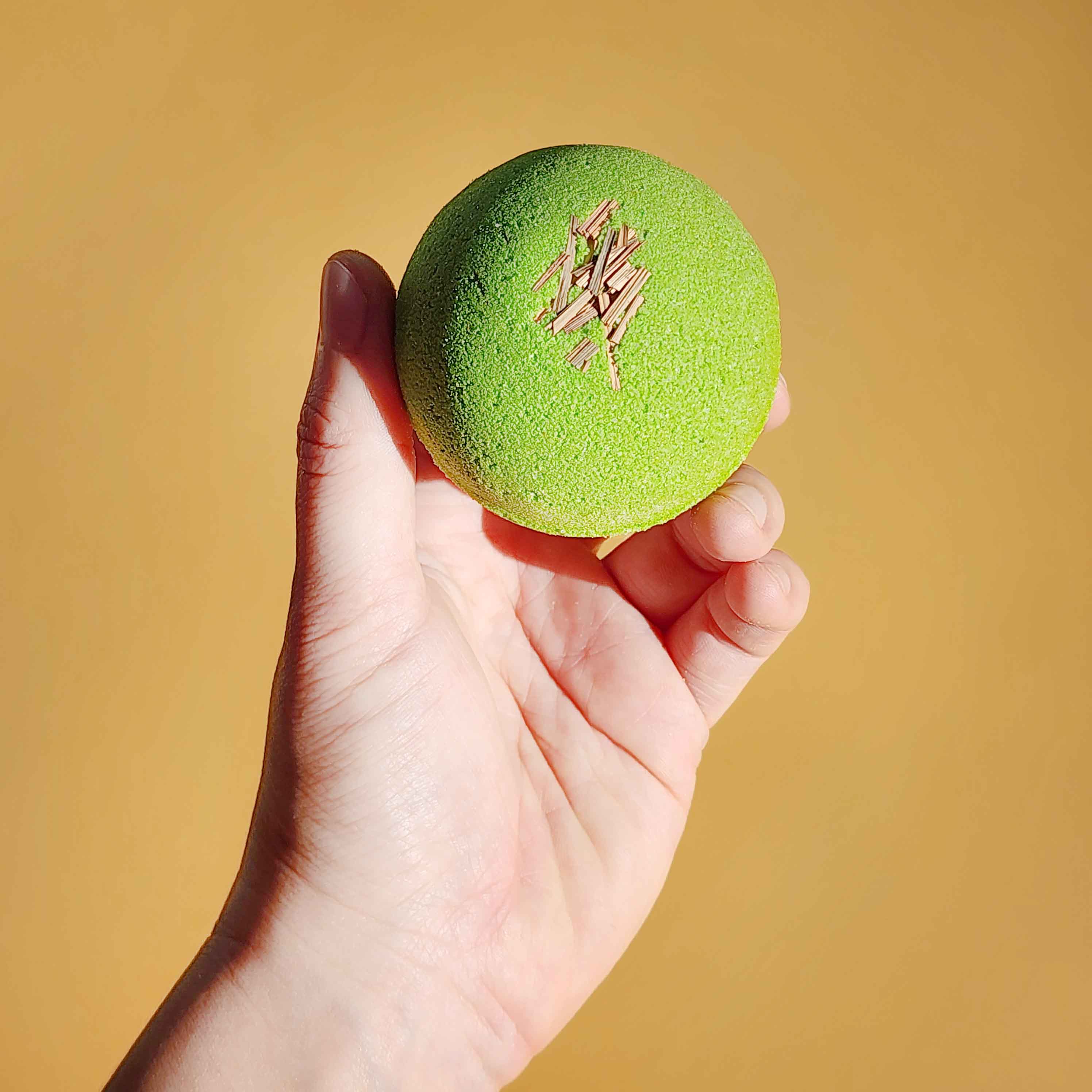 A hand holding a bright green Repair aromatherapy bath bomb topped with dried lemongrass against a yellow background