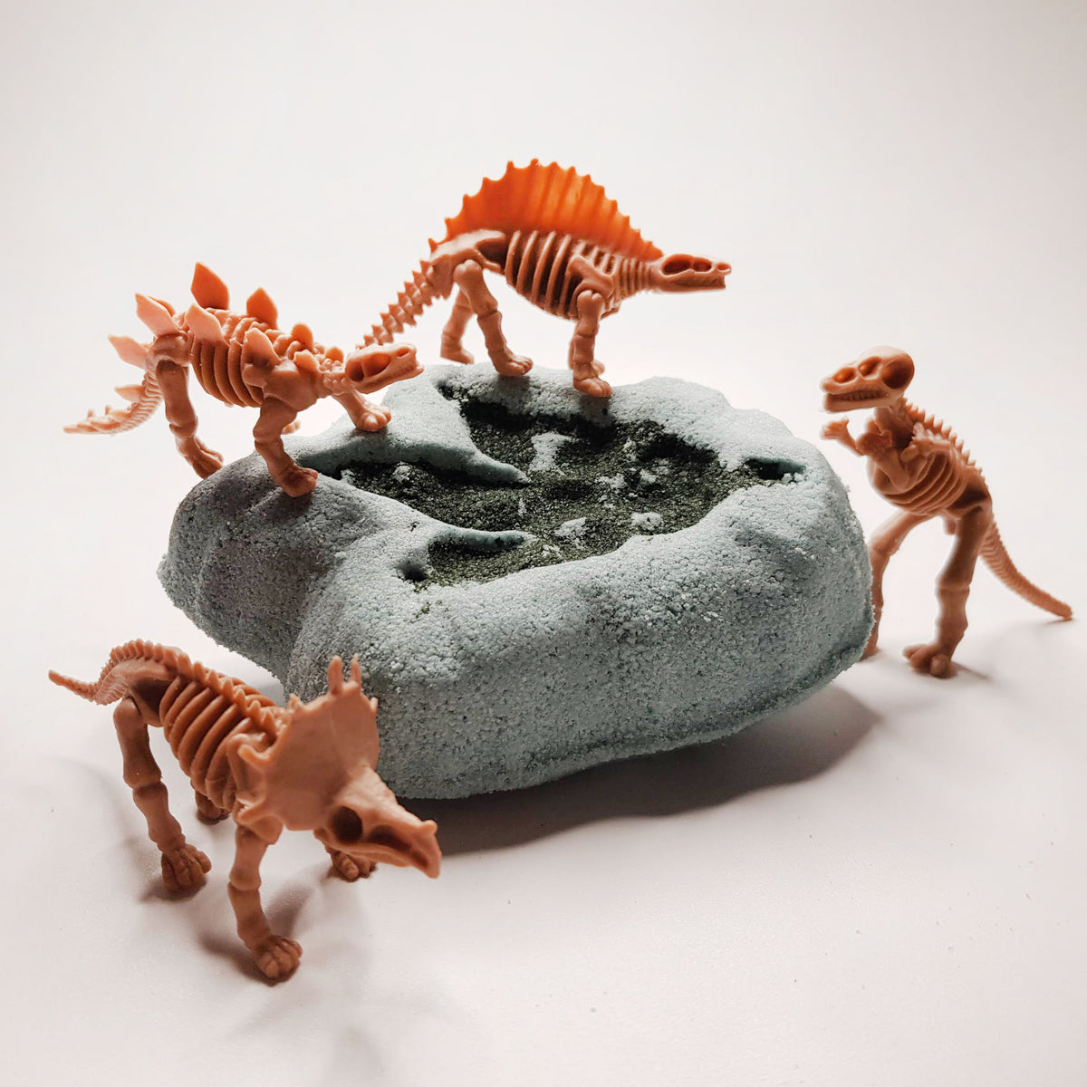 Grey bath bomb shaped like a dinosaur fossil with a dark dino footprint on it surrounded by skeletal dino toys