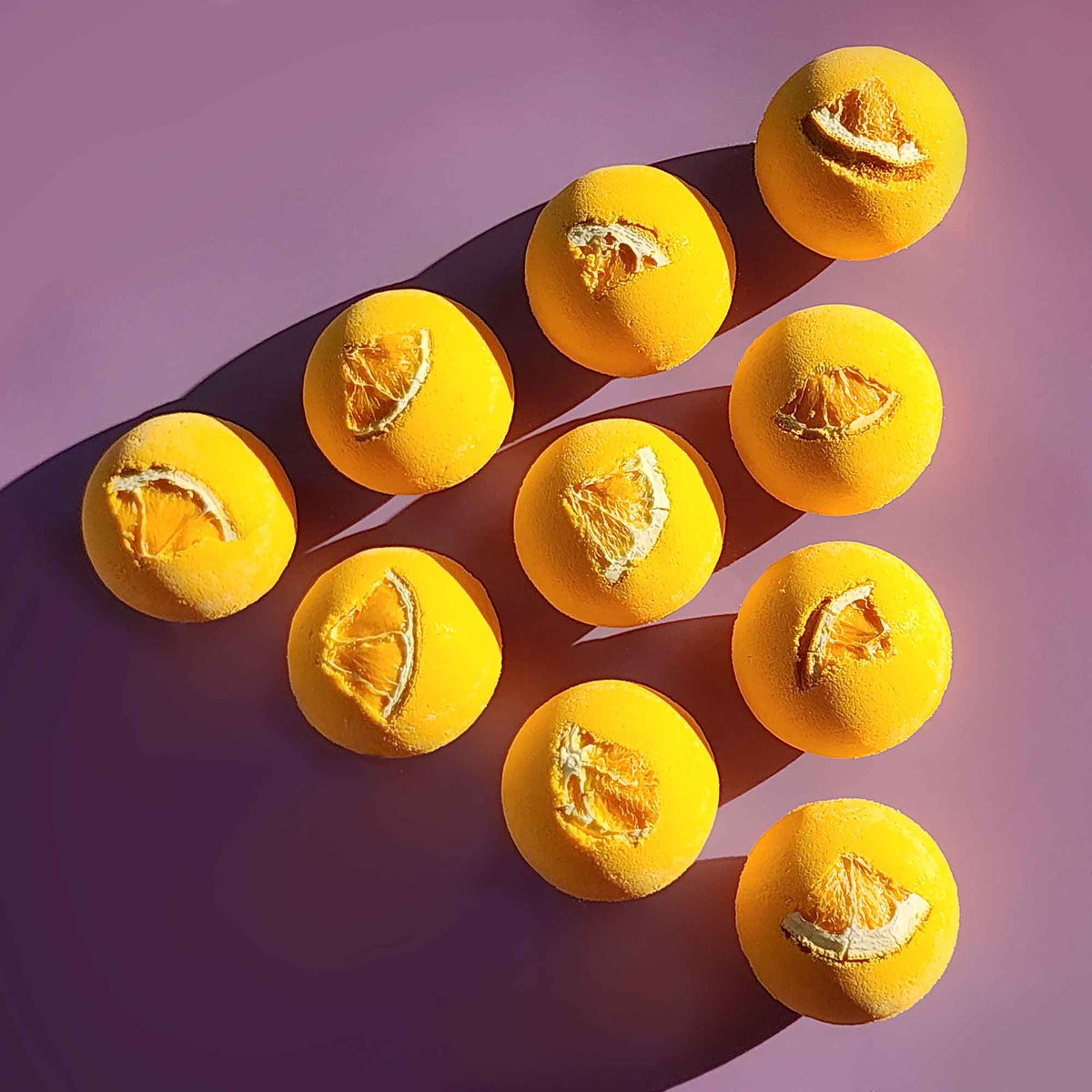 Ten yellow Energise aromatherapy bath bombs topped with dried orange slices arranged diagonally on a purple surface