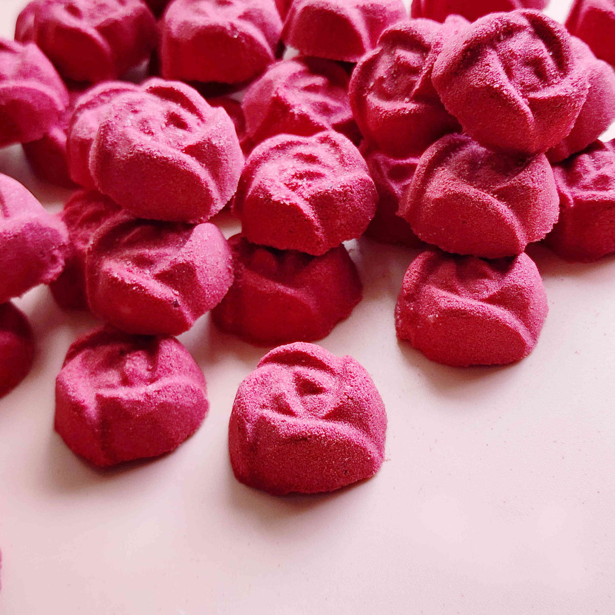 rose shaped mini bath bombs coloured a deep red laid out neatly on a plain pink background