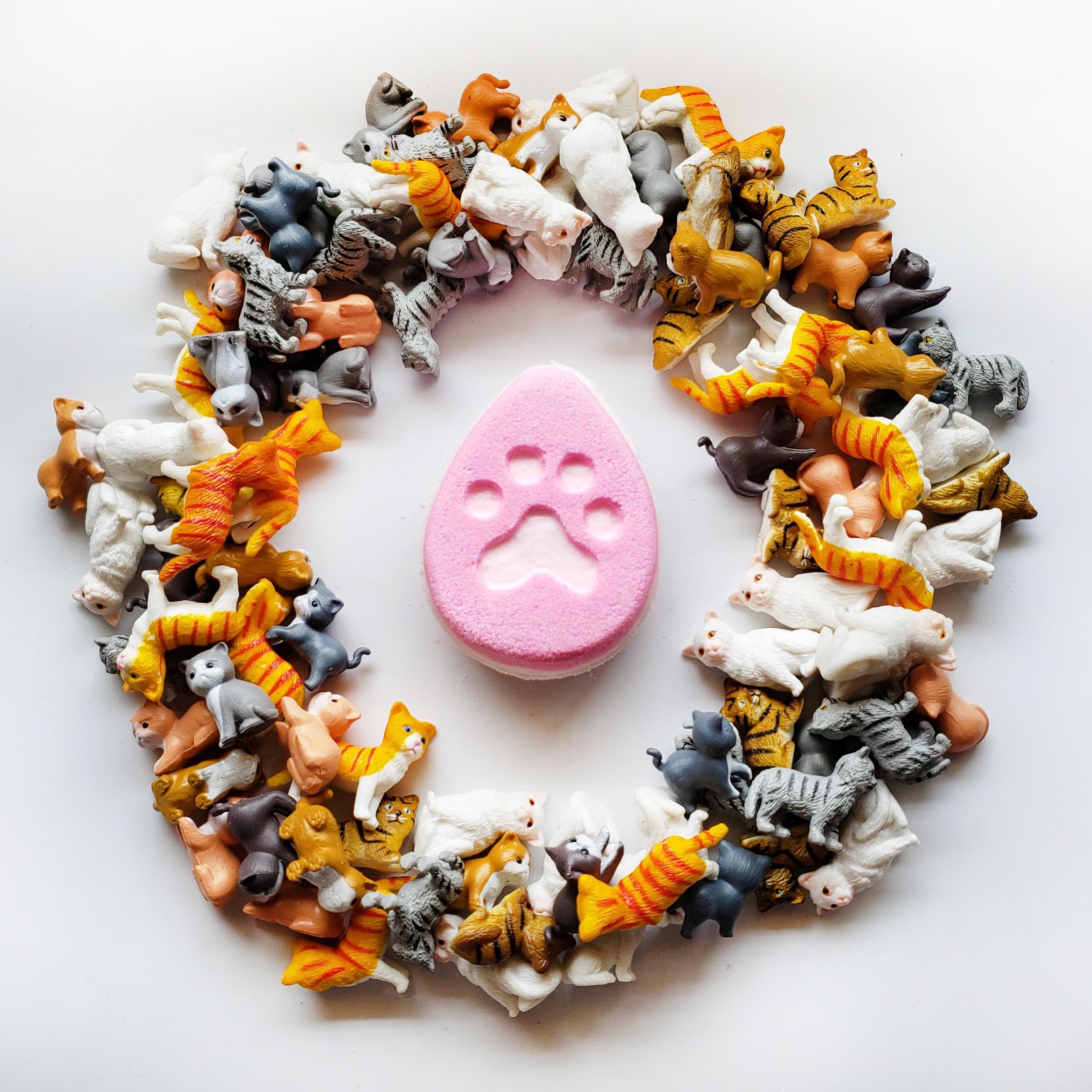 pink fat egg shaped bath bomb with a cat paw print embossed in the centre surrounded by miniature cat figurines