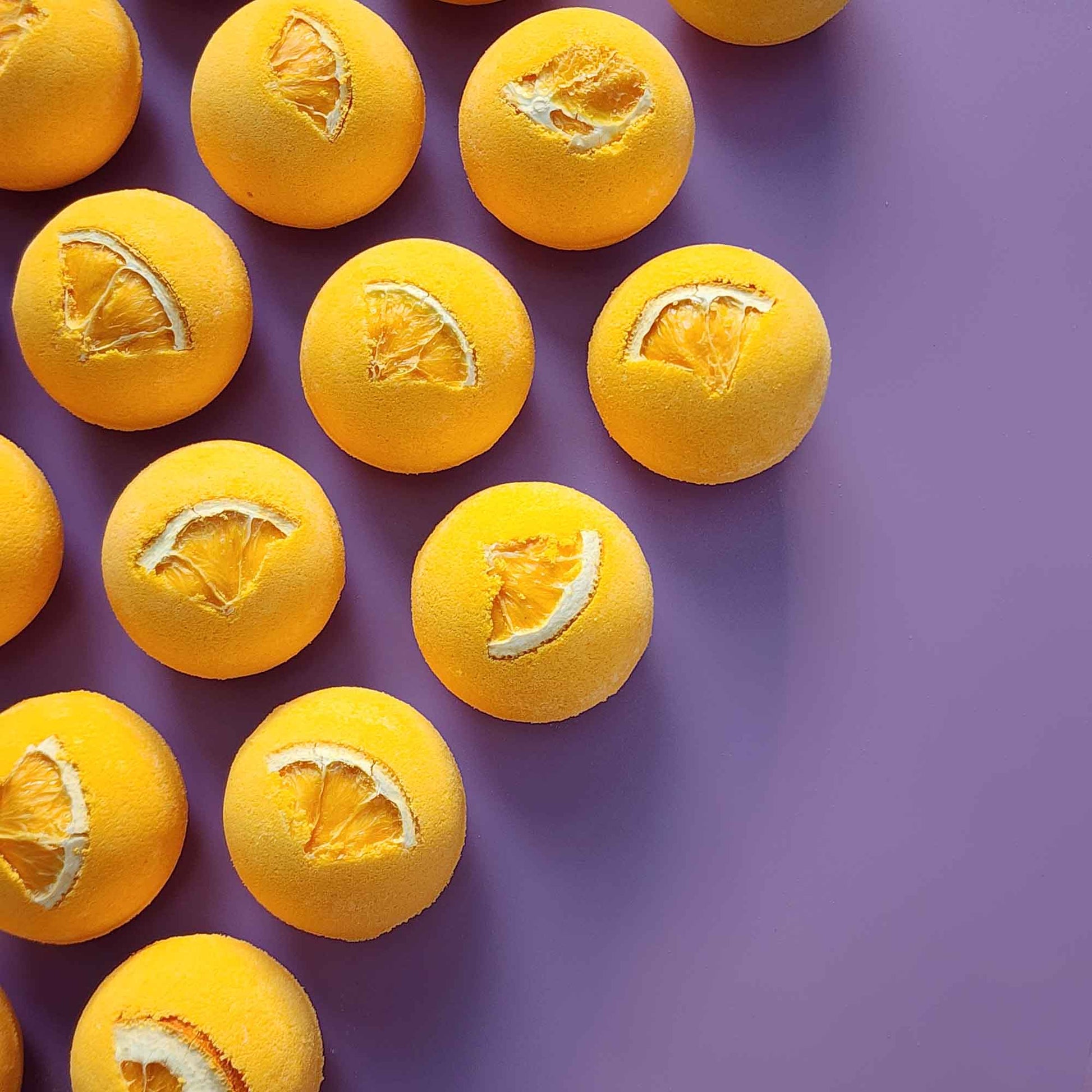 Top-down view of multiple yellow Energise aromatherapy bath bombs topped with dried orange slices on a purple background