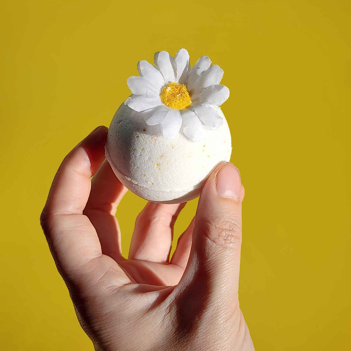 Hand holding a white chamomile bath bomb with a daisy flower topper against a yellow backdrop