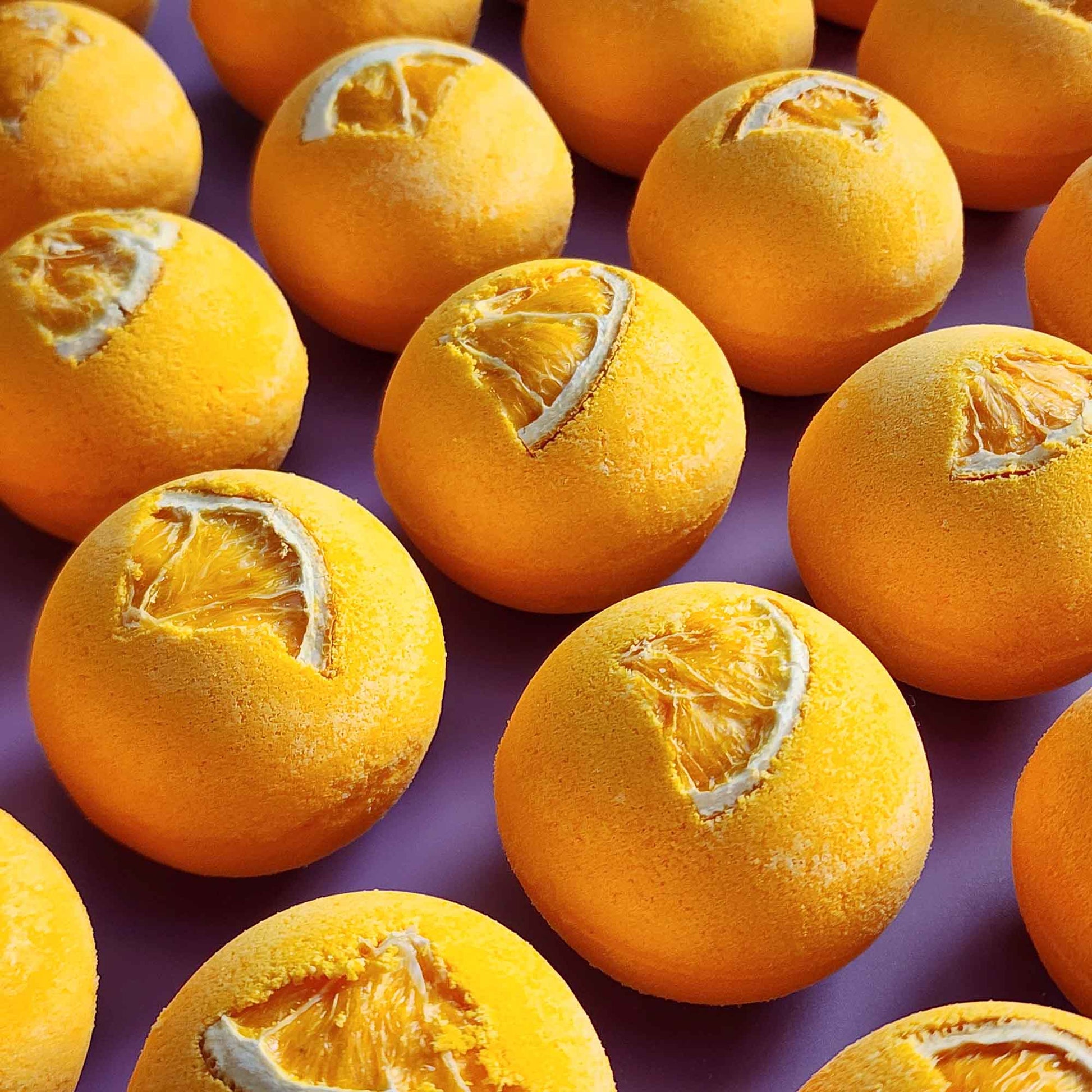 Top-down view of multiple yellow Energise aromatherapy bath bombs topped with dried orange slices on a purple background