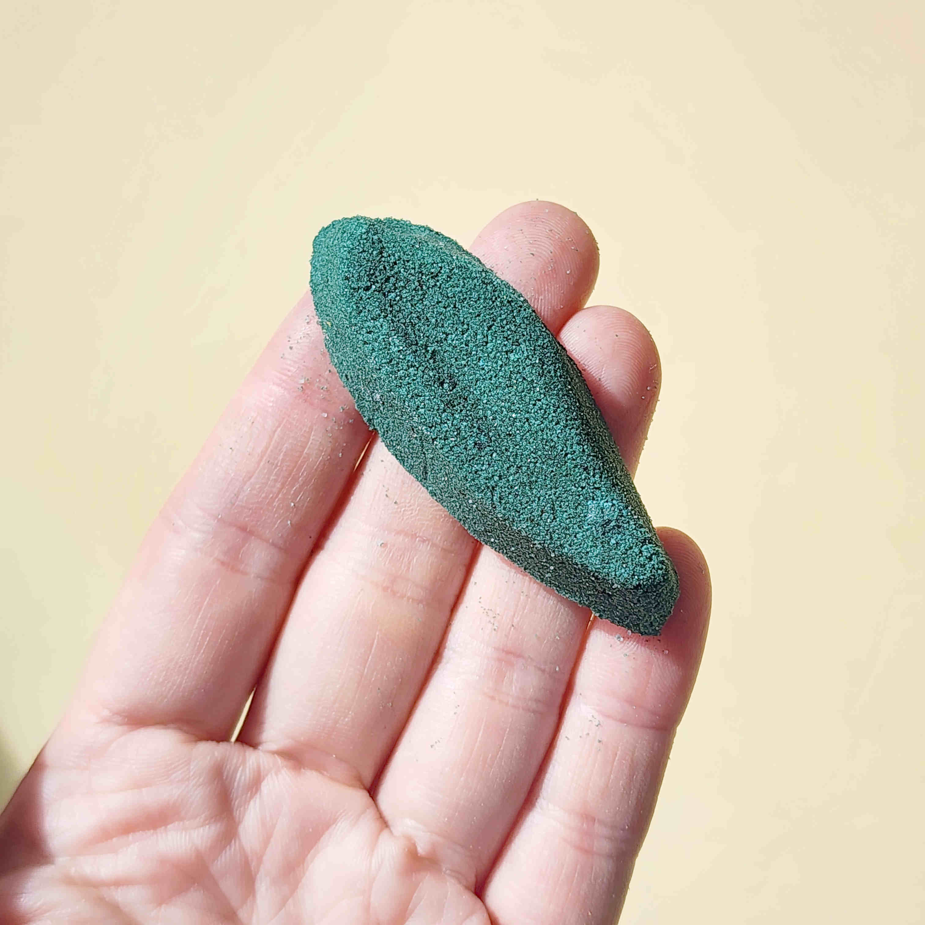 deep green long leaf shaped bath bomb held in a hand over a plain beige background