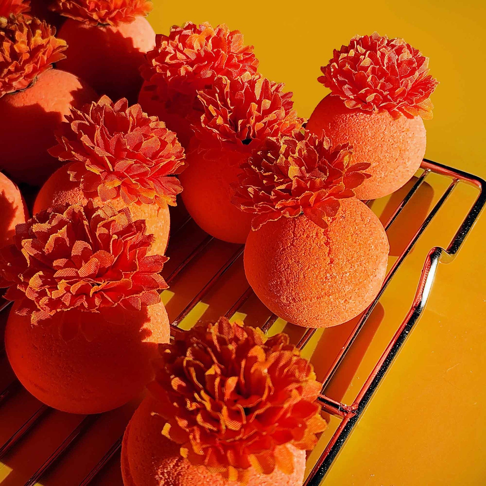Bright orange bath bombs topped with faux calendula flowers drying on a metal rack under warm sunlight.