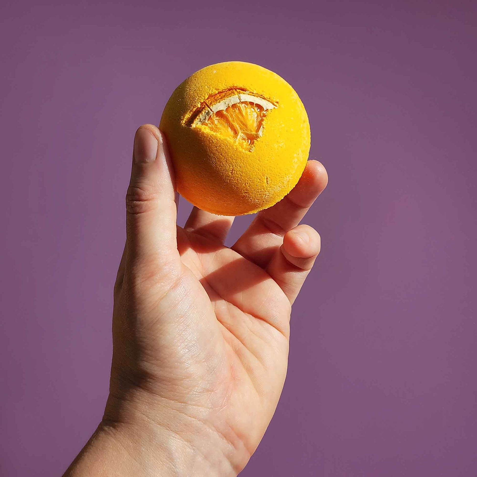 A hand holding a yellow Energise aromatherapy bath bomb topped with a dried orange slice against a purple background