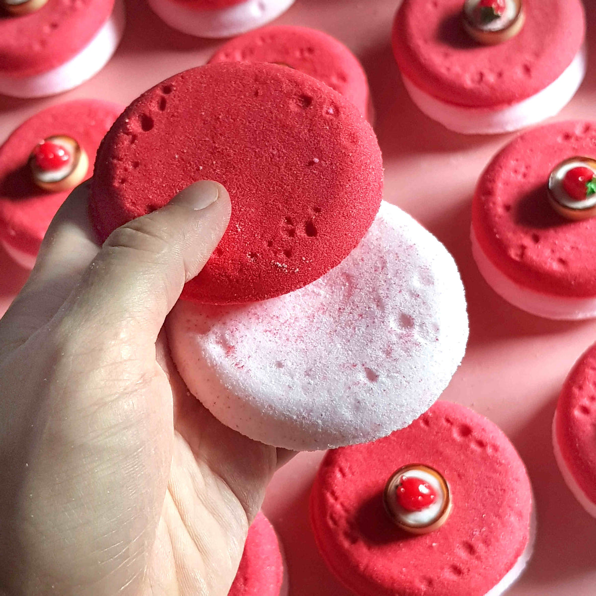 Flat disc shaped bath bombs stacked and placed in a tidy array on a plain pink background with one held in a hand close up