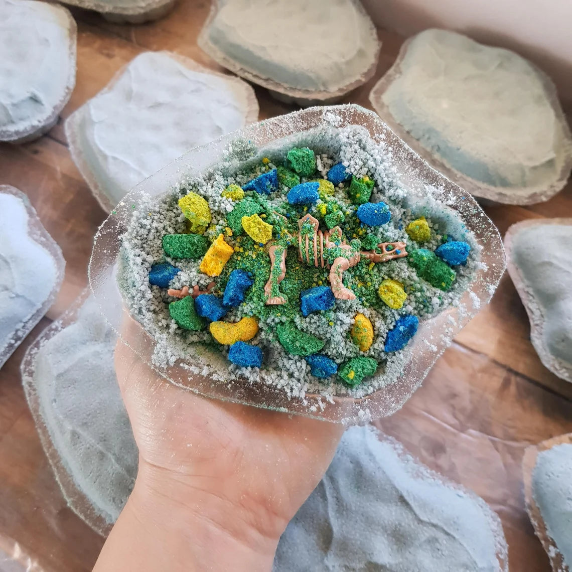 a partially completed bath bomb being held in a hand with lots of green, blue and yellow colour embeds
