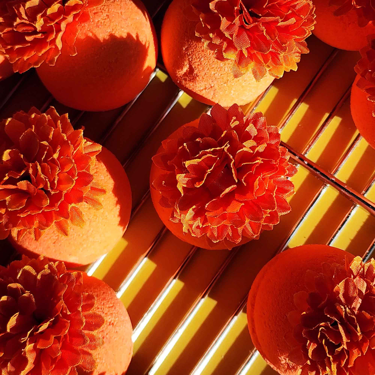 Bright orange bath bombs topped with faux calendula flowers drying on a metal rack under warm sunlight.