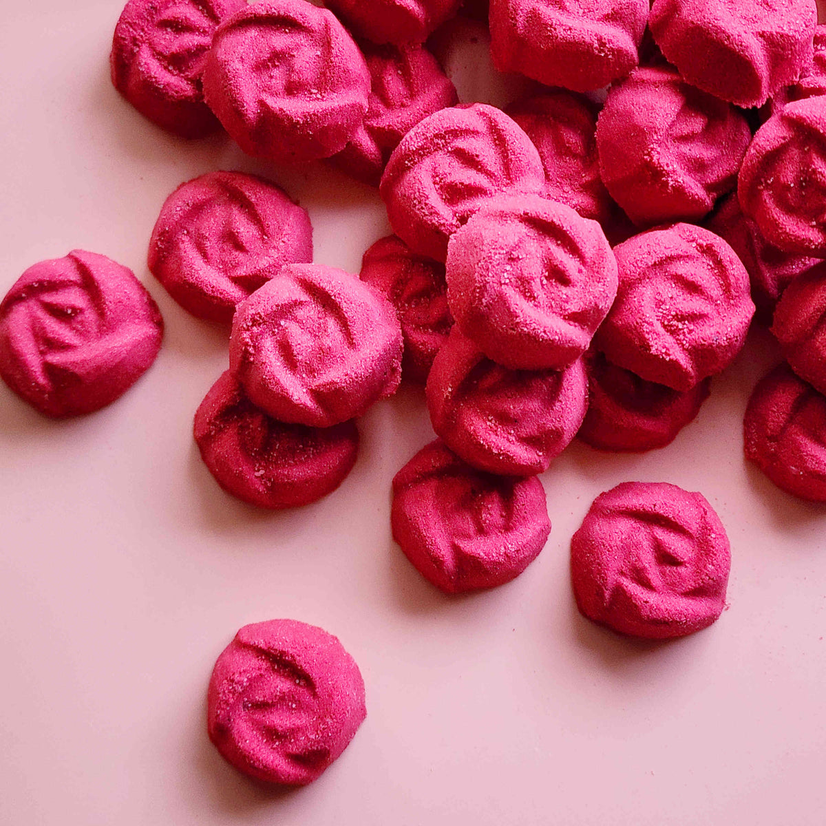 rose shaped mini bath bombs coloured a deep red laid out neatly on a plain pink background