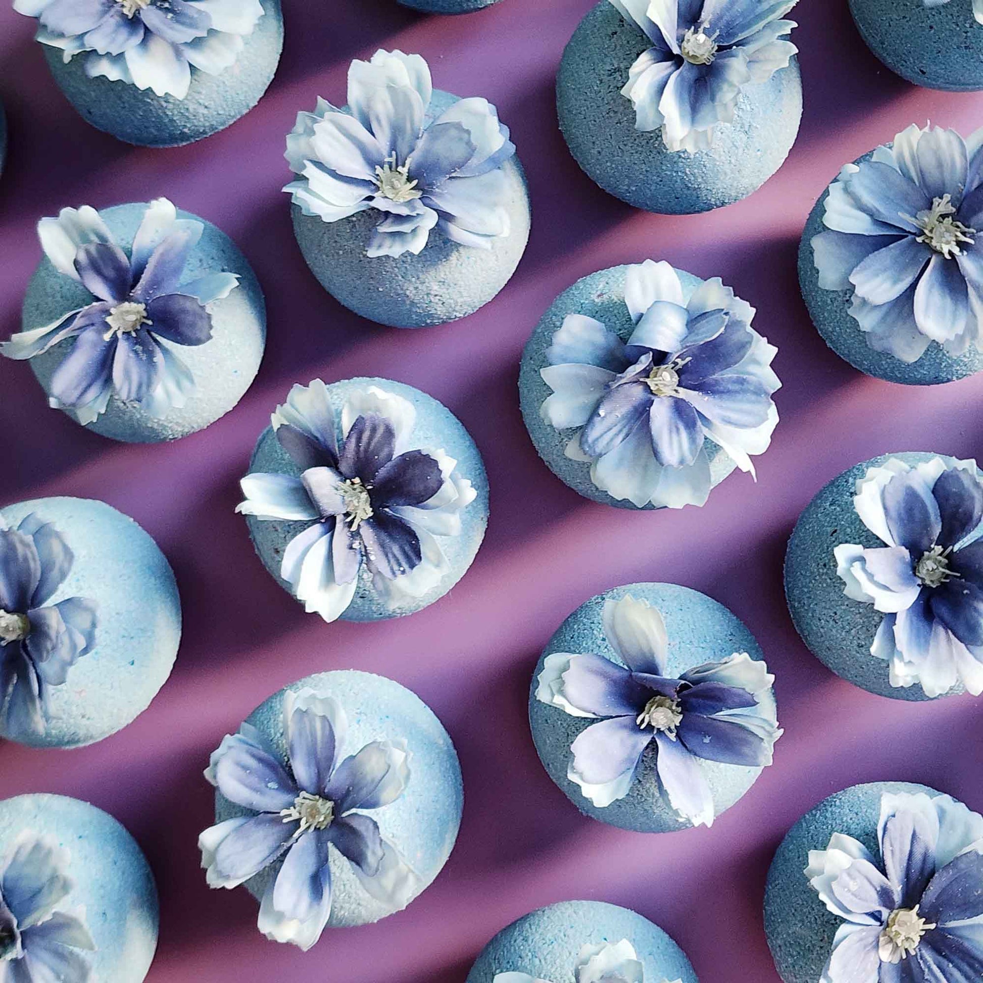 Overhead view of multiple blue wildflower bath bombs arranged neatly on a mauve surface, each topped with a faux blue flower