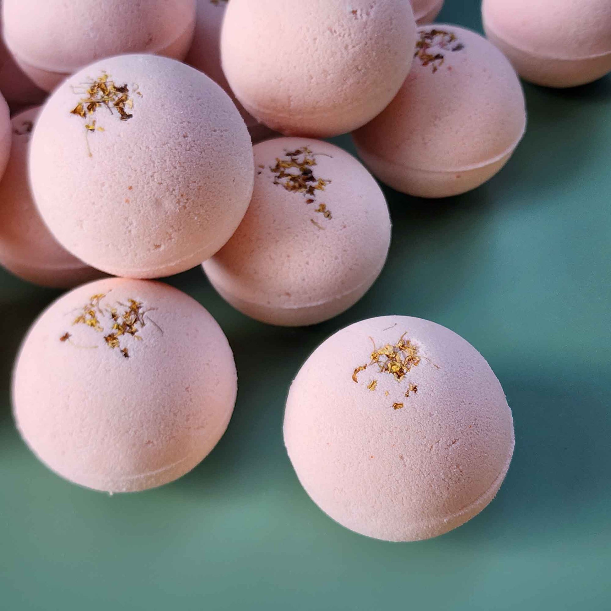 Multiple pale pink Grounding aromatherapy bath bombs topped with small dried osmanthus flowers, arranged on a soft green back