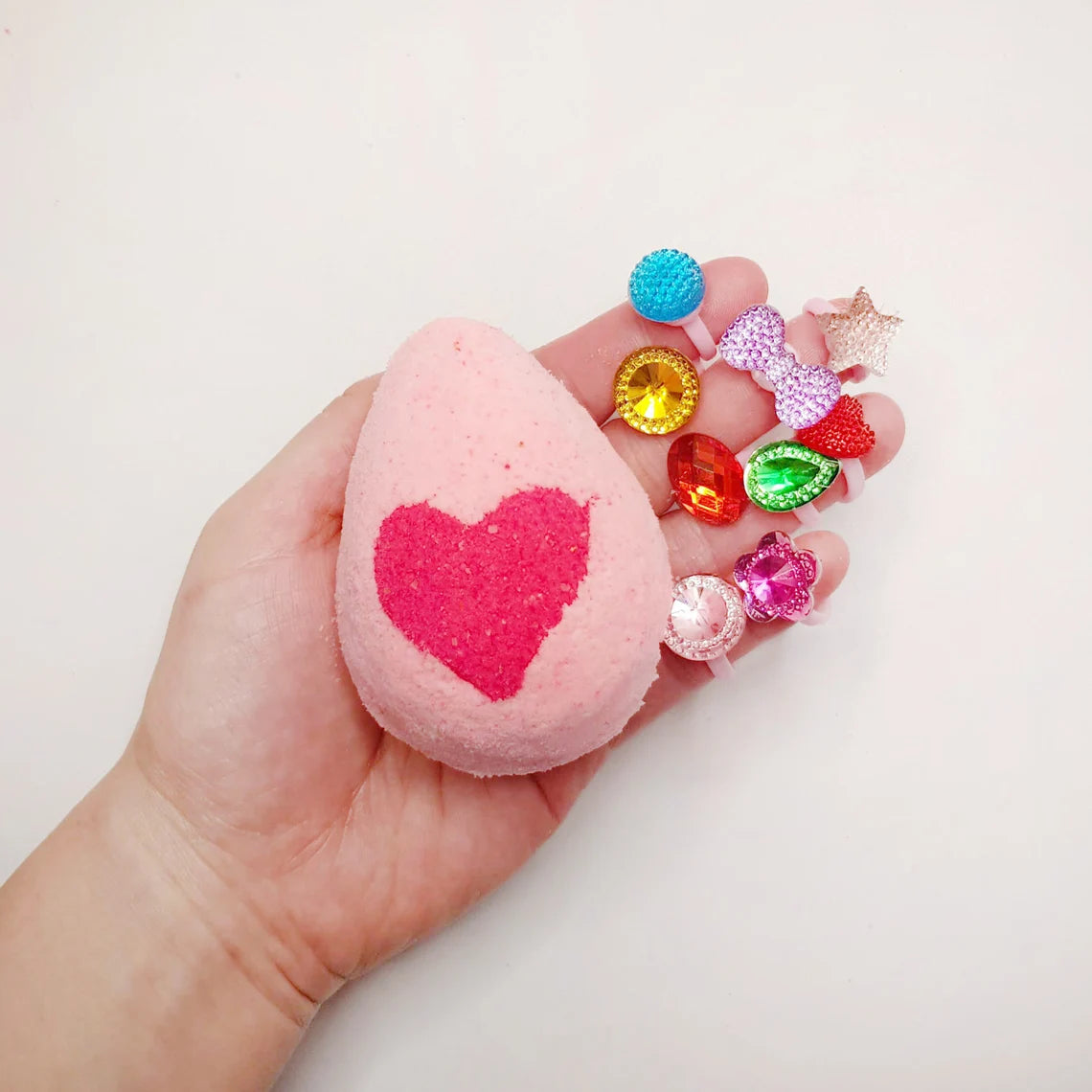 pink egg shaped bath bomb with a bright pink heart in the middle being held in a hand with toy rings