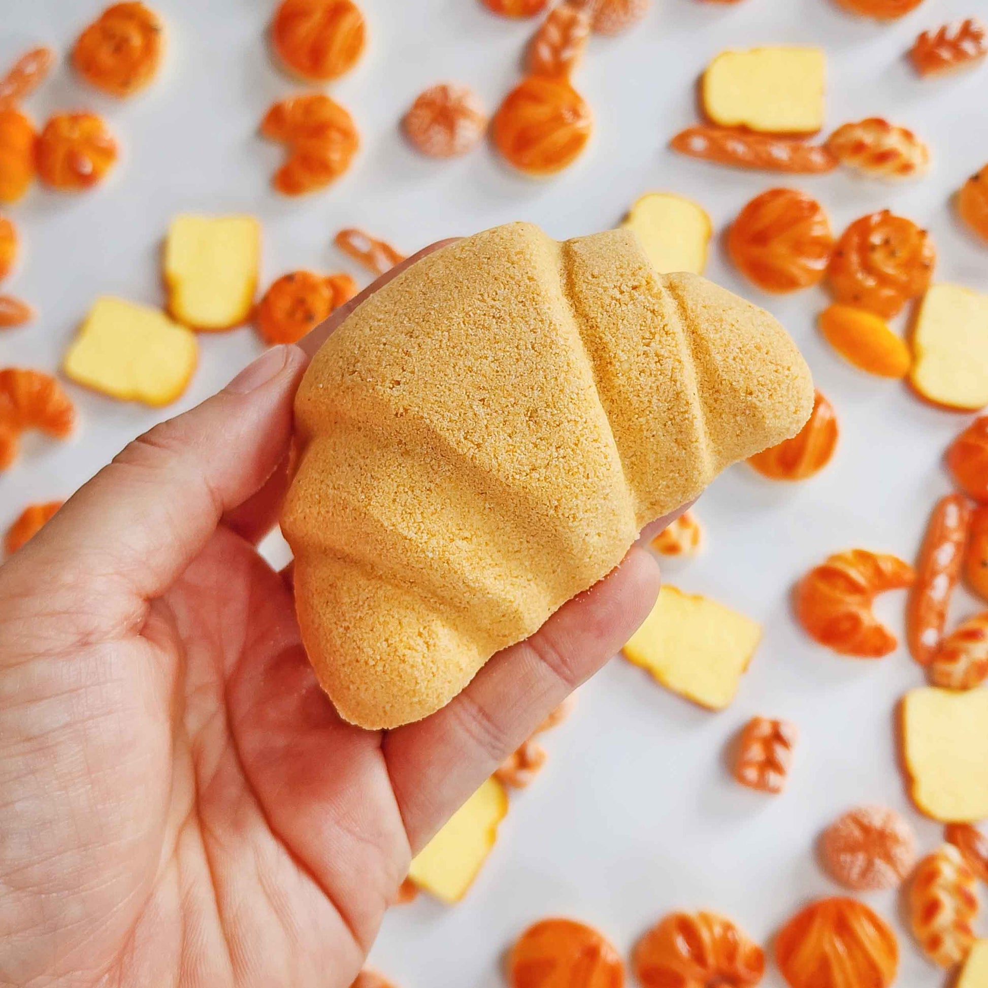 Hand holding a croissant-shaped bath bomb over miniature bread toys