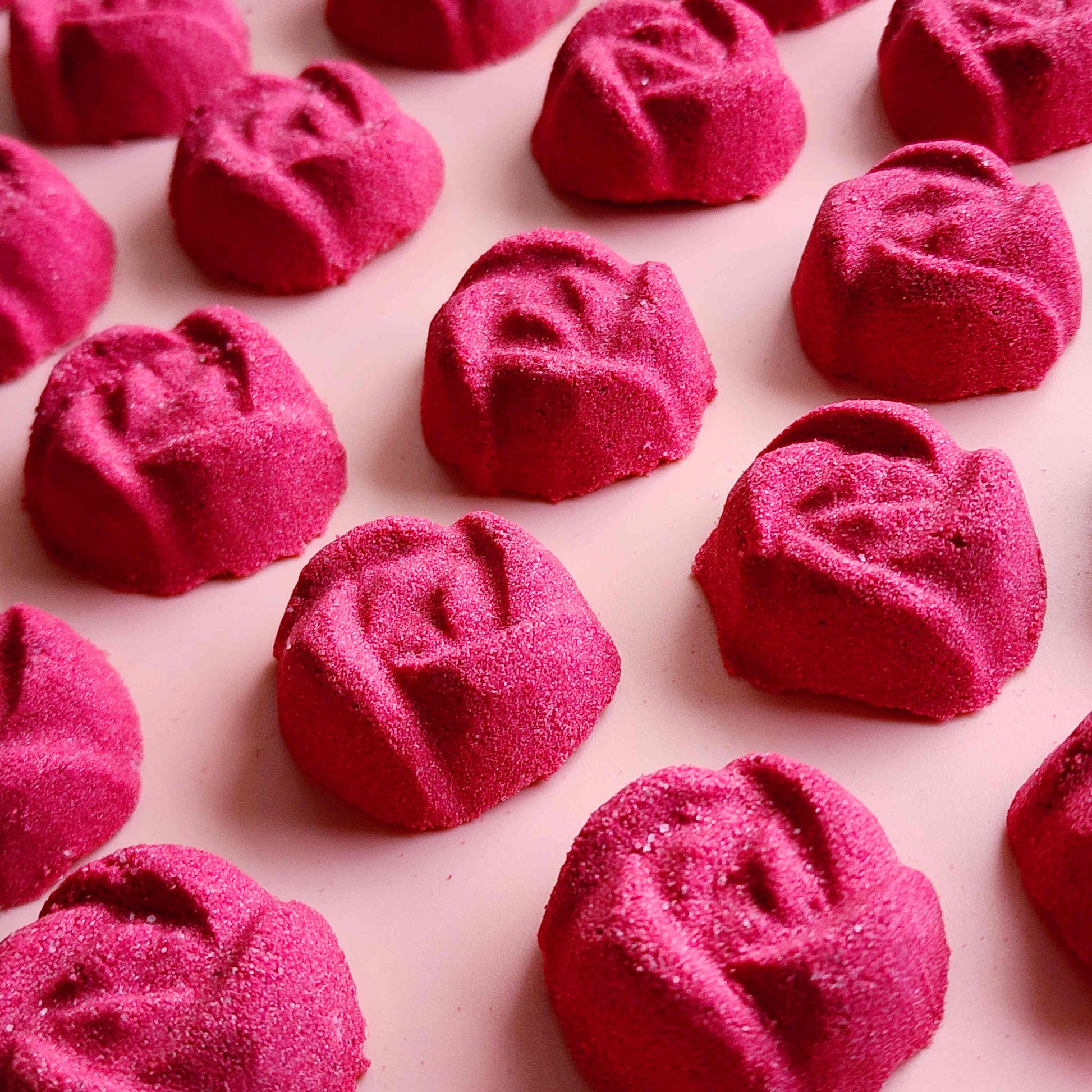 rose shaped mini bath bombs coloured a deep red laid out neatly on a plain pink background