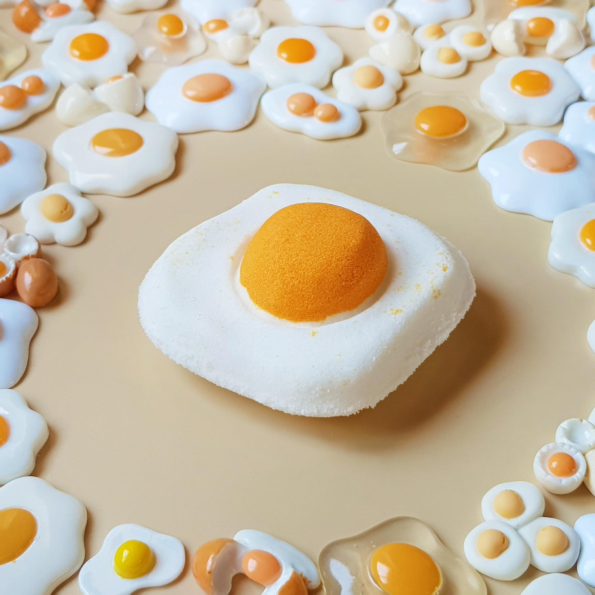 Sunny-side-up egg-shaped mango and cream bath bomb surrounded by miniature fried-egg decorations